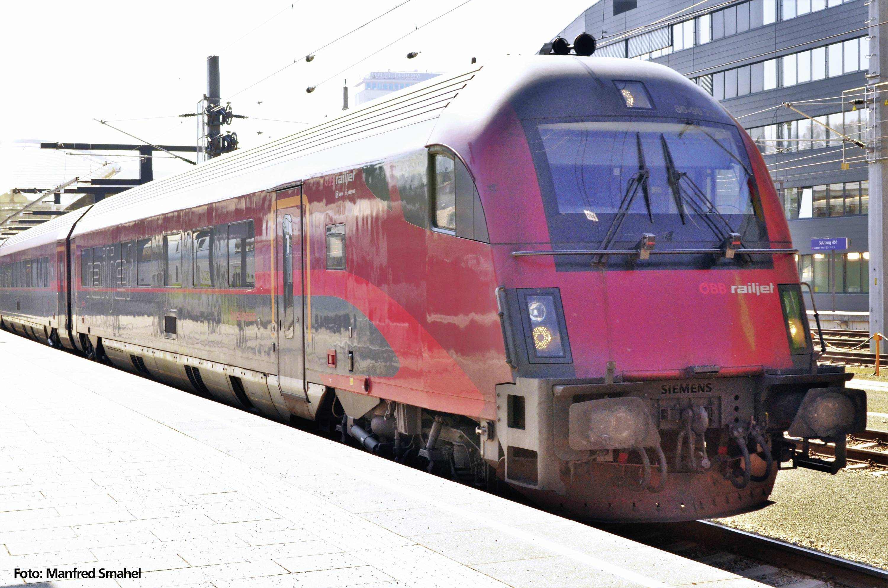Ein roter Zug der ÖBB Railjet steht an einem Bahnsteig bei sonnigem Wetter. Er ist modern gestaltet und bereit zur Abfahrt.