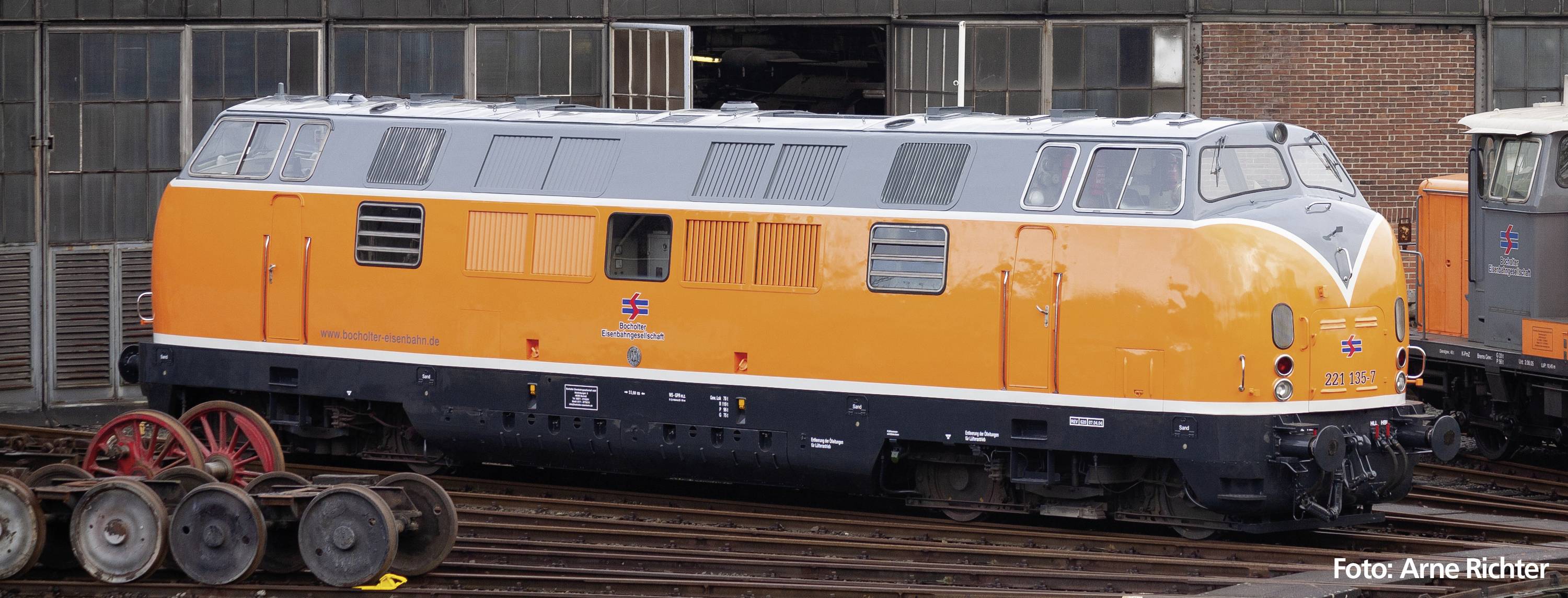 Eine orangefarbene Lokomotive steht vor einem Bahndepot. Sie hat ein markantes Design und gehört zu einer historischen Zugflotte.