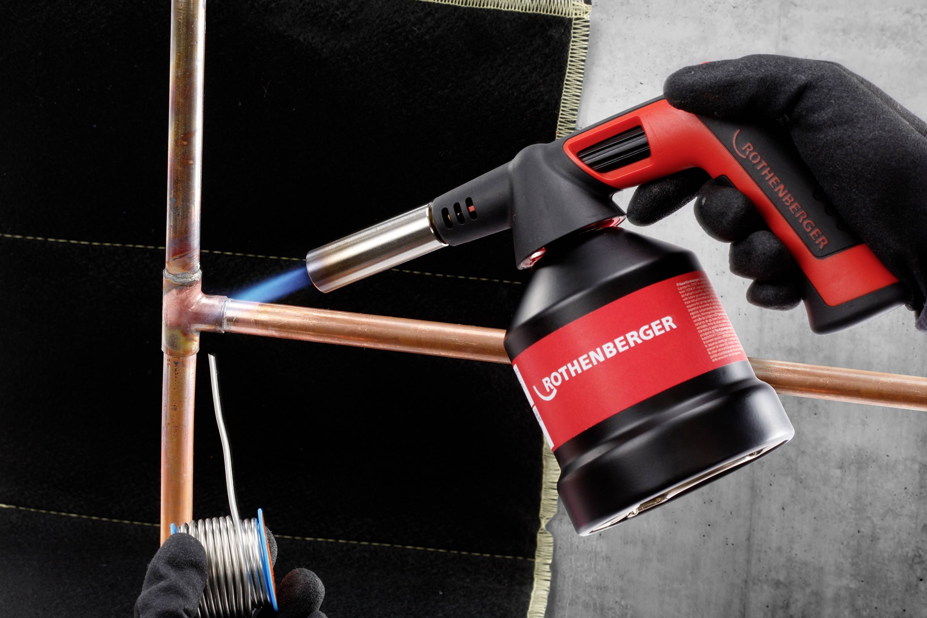 Eine Person lötet Kupferrohre mit einem Rothenberger Lötkolben, Flamme sichtbar. Werkzeug und Hände im Bild.