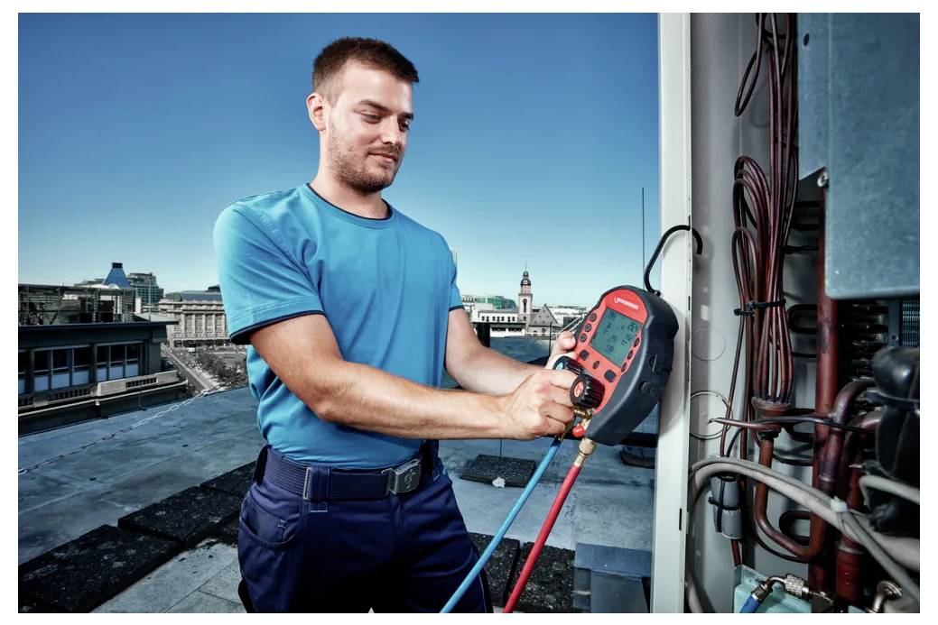 Ein Techniker in einem blauen Hemd nutzt ein digitales Multimeter, um HVAC-Ausrüstung auf einem Dach zu überprüfen. Eine Stadtlandschaft ist im Hintergrund sichtbar.