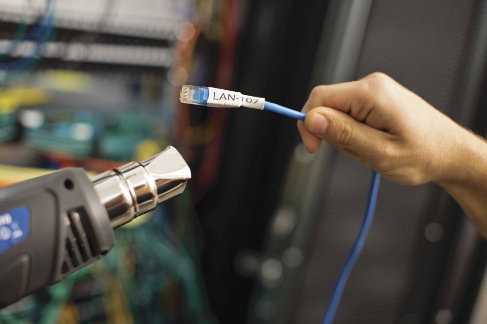 Eine Hand hält ein beschriftetes LAN-Kabel vor einen Föhn. Im Hintergrund sind unscharfe Computerkabel zu sehen.