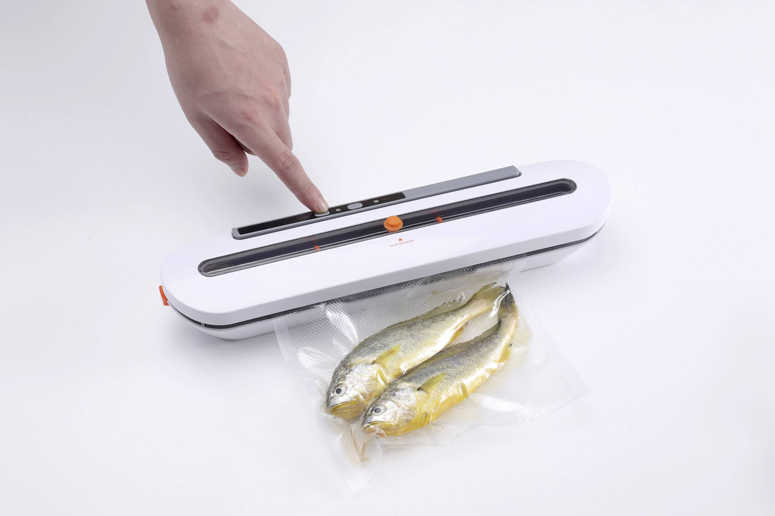 Eine Hand bedient ein Folienschweißgerät, das zwei vakuumverpackte Fische auf einem weißen Hintergrund versiegelt.