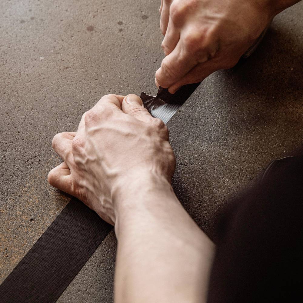 Eine Person befestigt ein schwarzes Klebeband auf einer Oberfläche. Ihre Hände sind sichtbar und konzentriert, um das Band sorgfältig anzubringen.