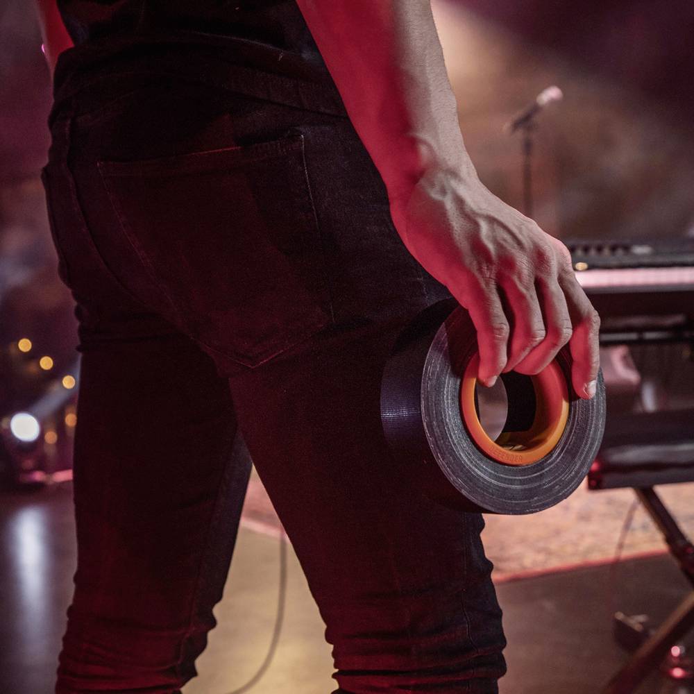 Eine Person hält Klebeband in der Hand, steht auf einer Bühne; im Hintergrund sind undeutlich ein Mikrofon und Musikgeräte erkennbar.