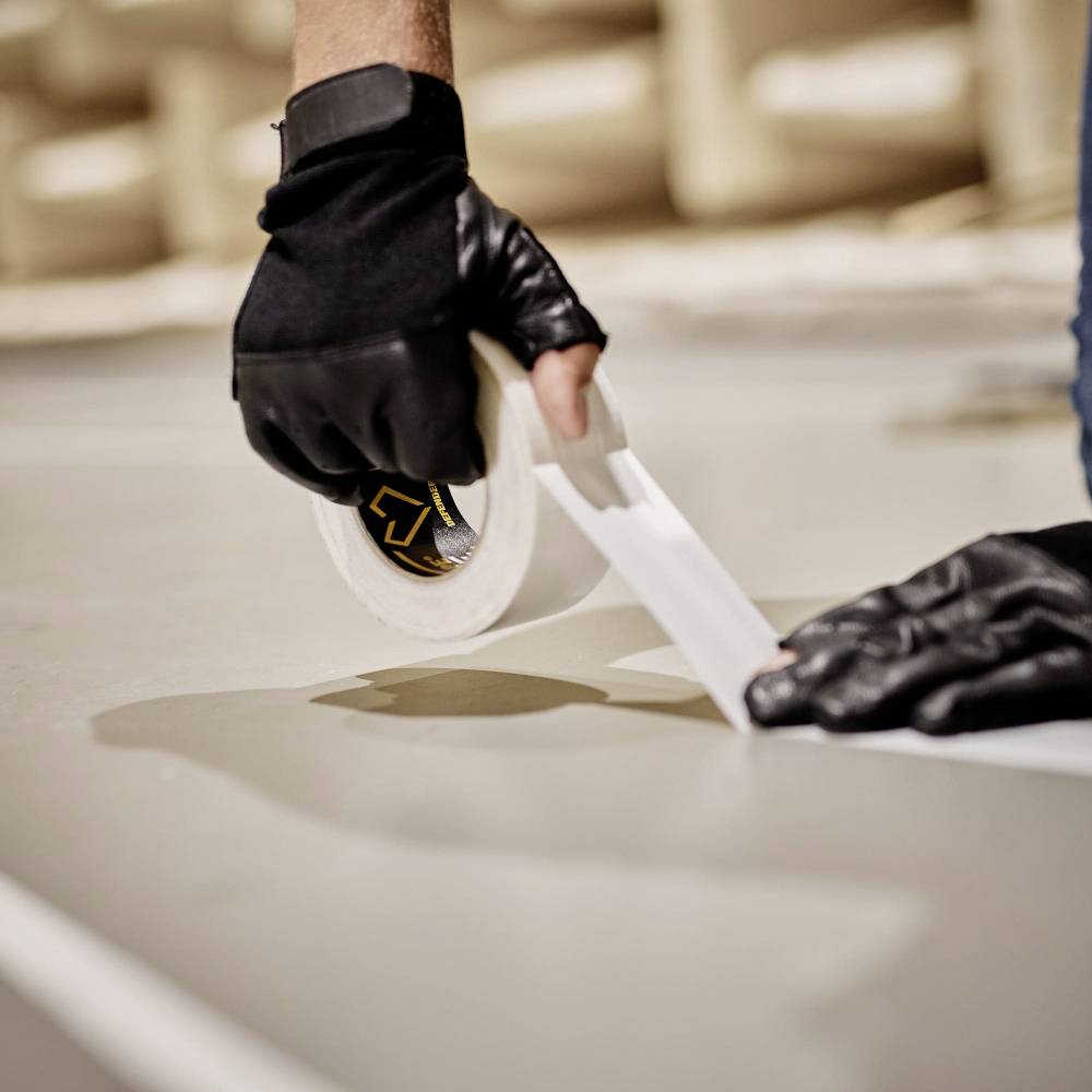 Eine Person mit Handschuhen klebt einen Streifen Klebeband auf eine glatte Oberfläche. Die Umgebung wirkt technisch und funktional.