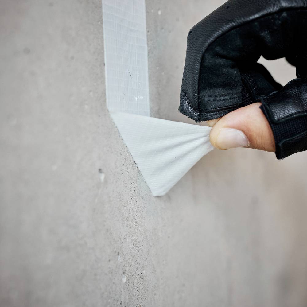 Eine Hand mit Handschuh zieht Klebeband von einer Betonwand ab.