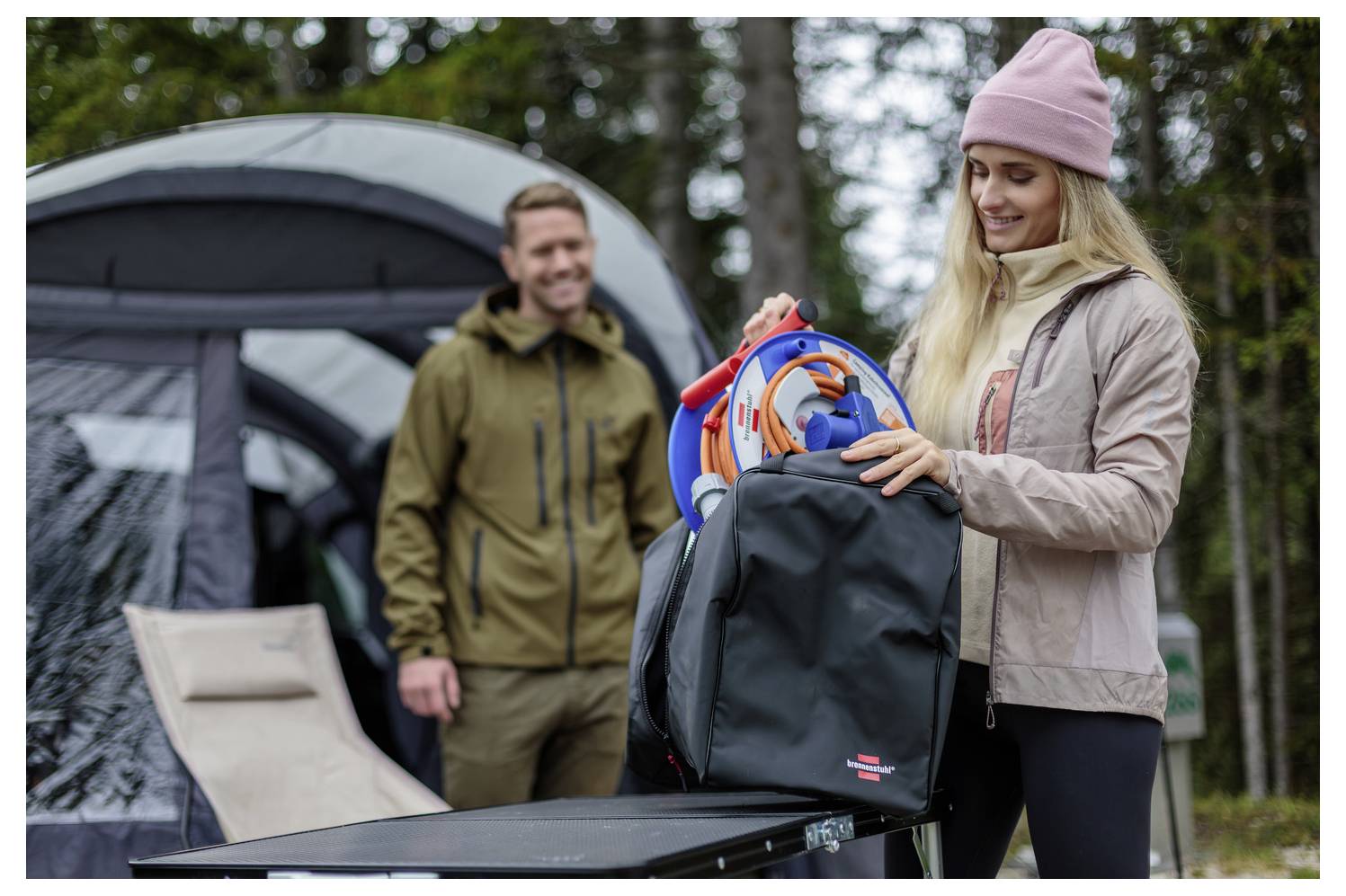 Frau lädt Campingausrüstung in einen Kofferraum eines Autos auf einem Campingplatz, mit einem Mann und einem Zelt im Hintergrund.