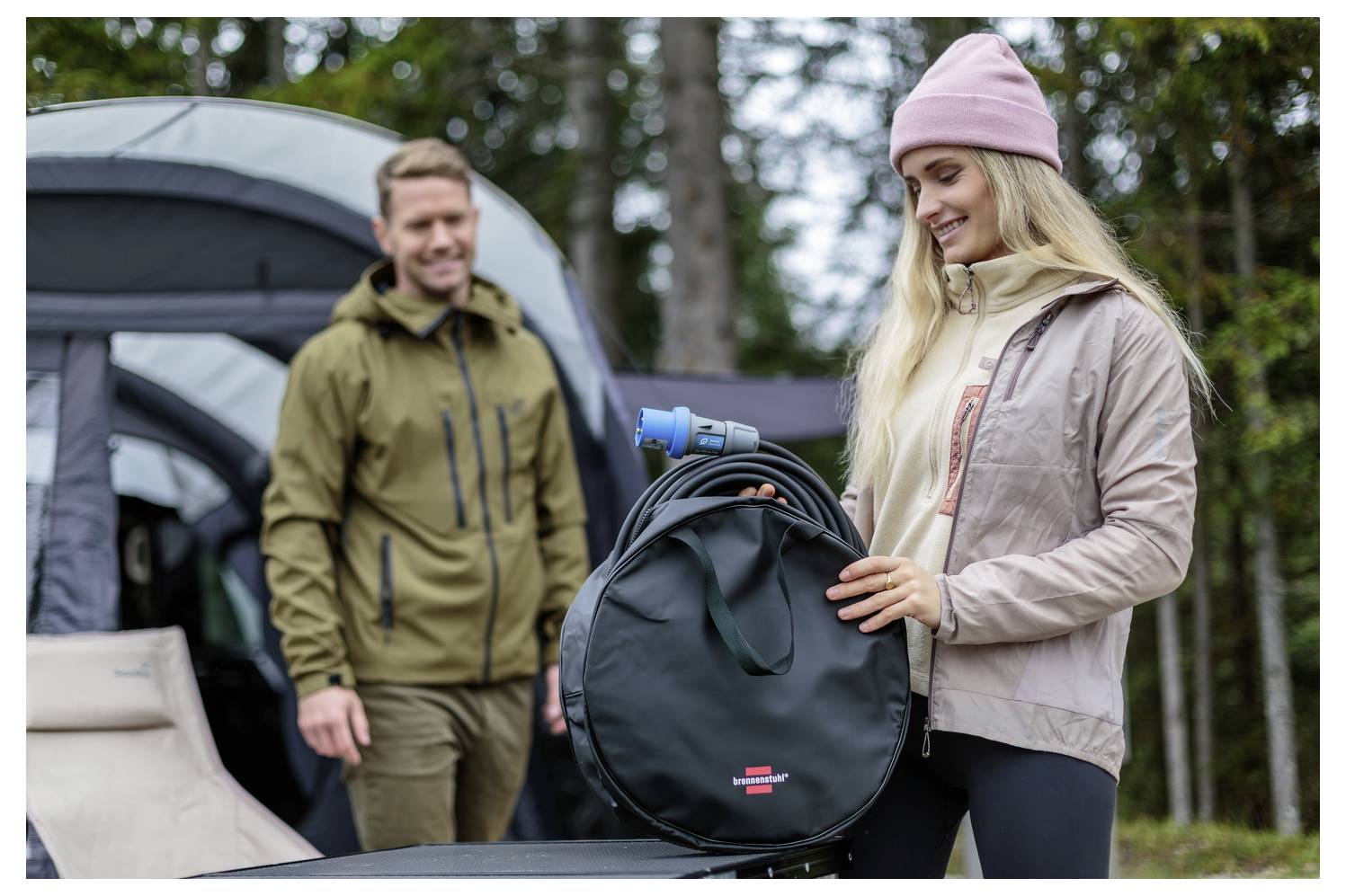 Frau an einem Campingplatz hält eine schwarze runde Tasche mit einem blauen Campingkocher, während ein Mann im Hintergrund in der Nähe eines Zelts steht.