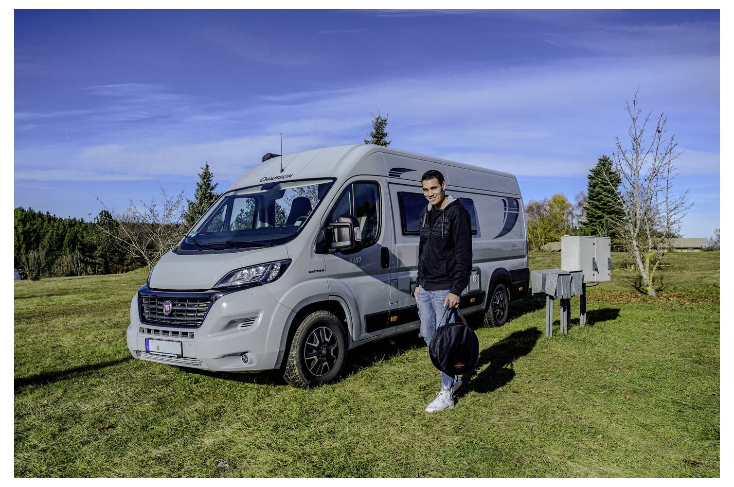 Ein Mann steht neben einem silbernen Wohnmobil, das auf einer Wiese unter einem blauen Himmel geparkt ist.