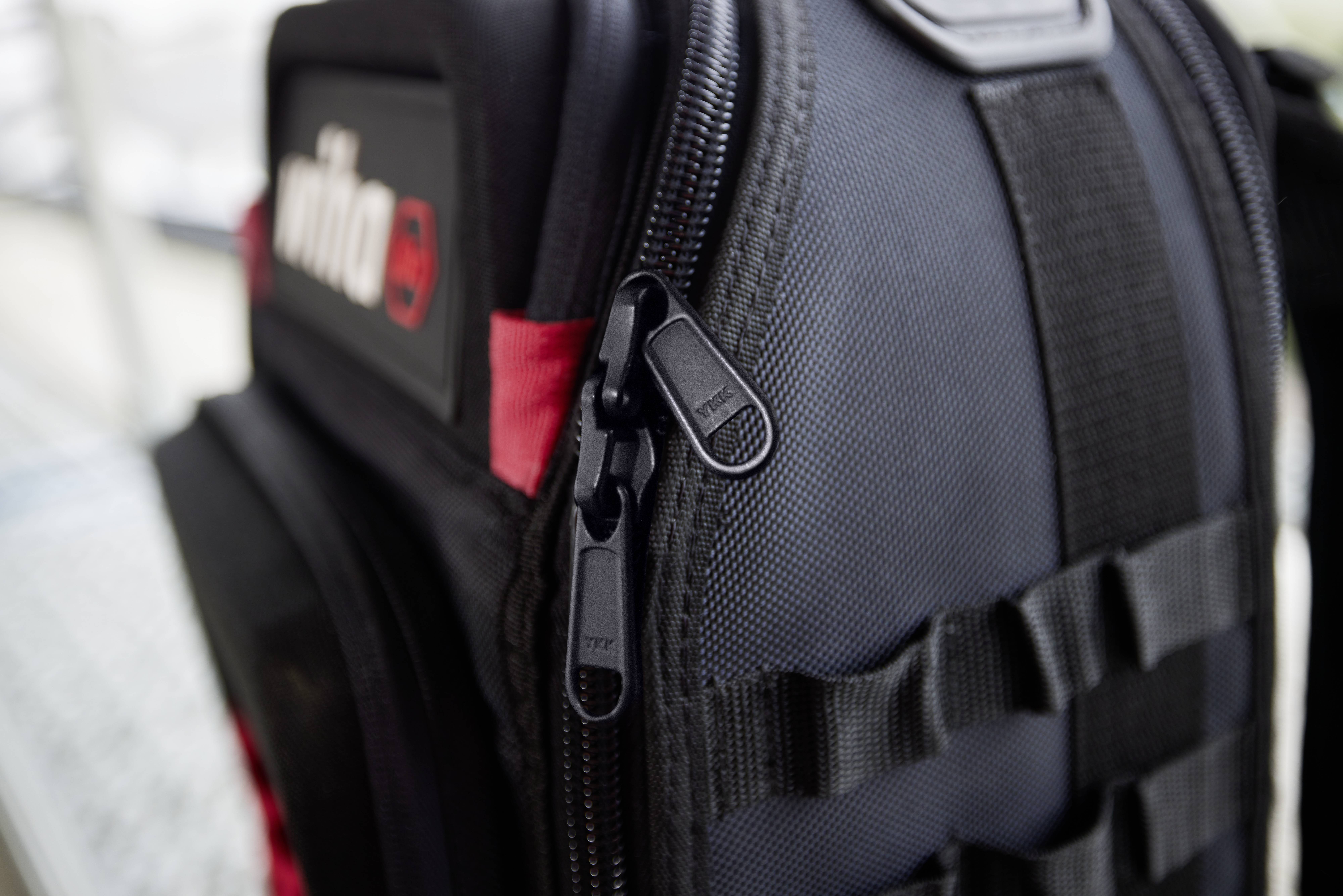 Ein schwarzer Rucksack mit einem Reißverschluss ist in Nahaufnahme zu sehen, mit einem roten Logo auf der linken Seite.