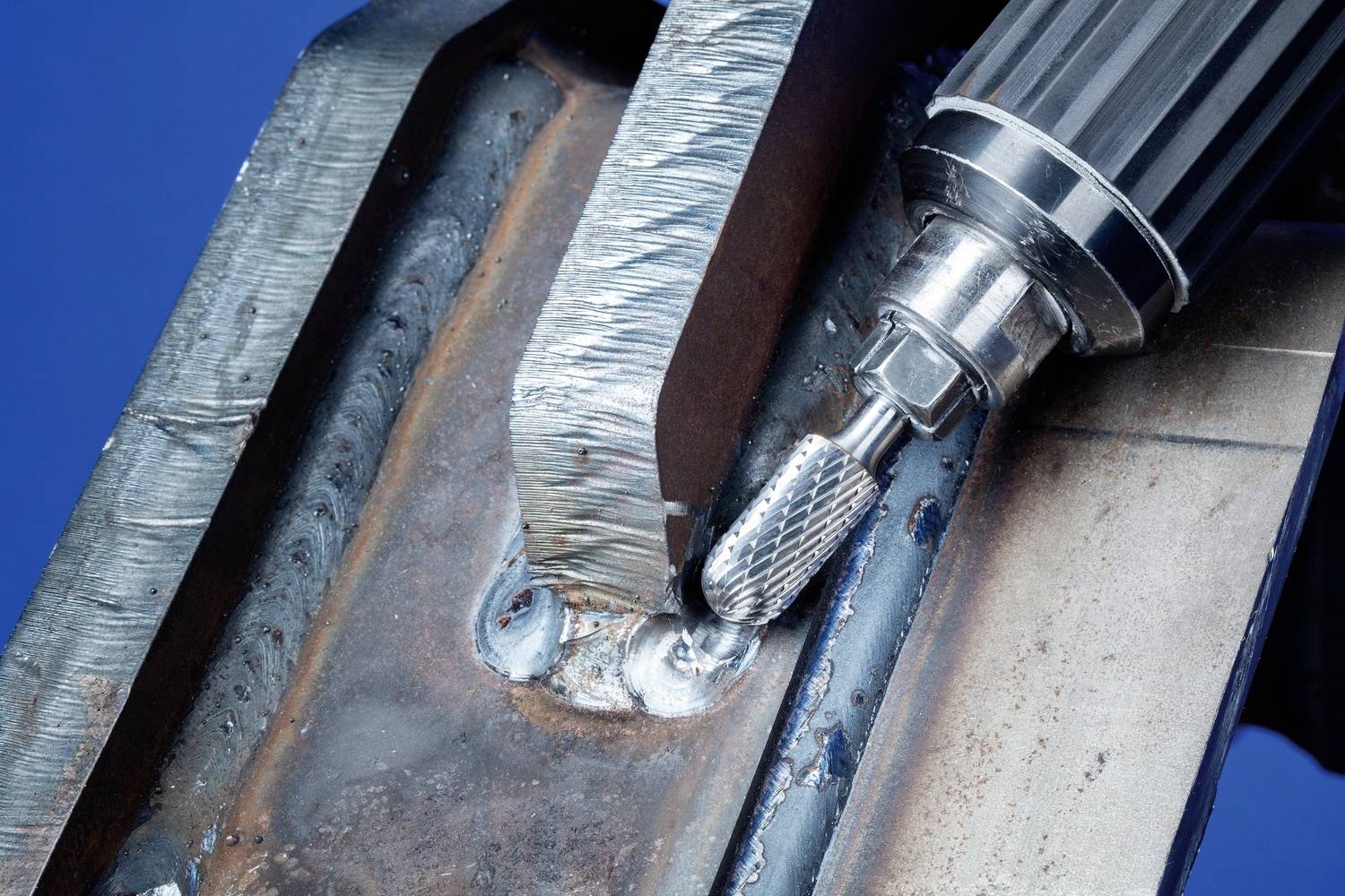 Nahaufnahme eines Metallbohrers, der eine Schweißnaht auf einer Metallplatte bearbeitet. Der Hintergrund ist blau.