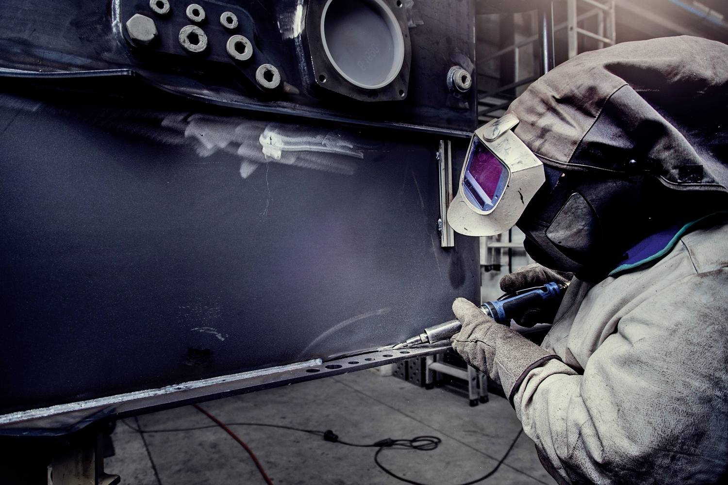 Ein Schweißer in Schutzkleidung arbeitet mit einem Schweißgerät an einem Metallstück in einer Werkstatt. Hintergrund zeigt industrielle Umgebung.