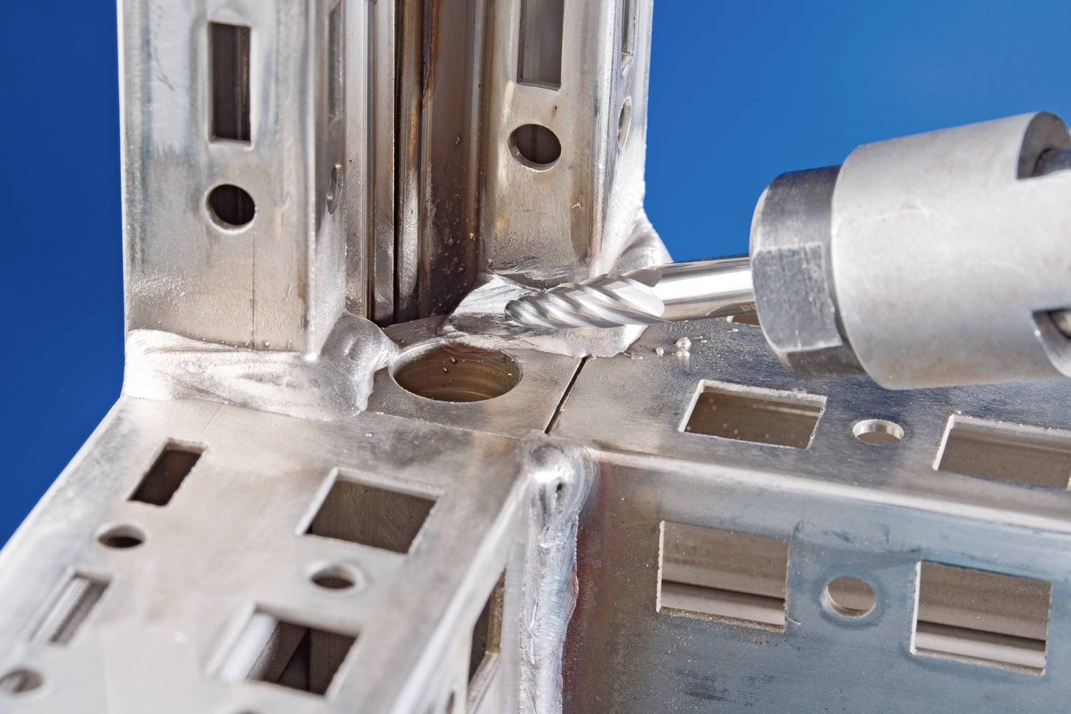 Ein Metallbohrer bearbeitet ein präzise gefertigtes Metallstück mit mehreren Löchern und Schlitzen vor blauem Hintergrund.