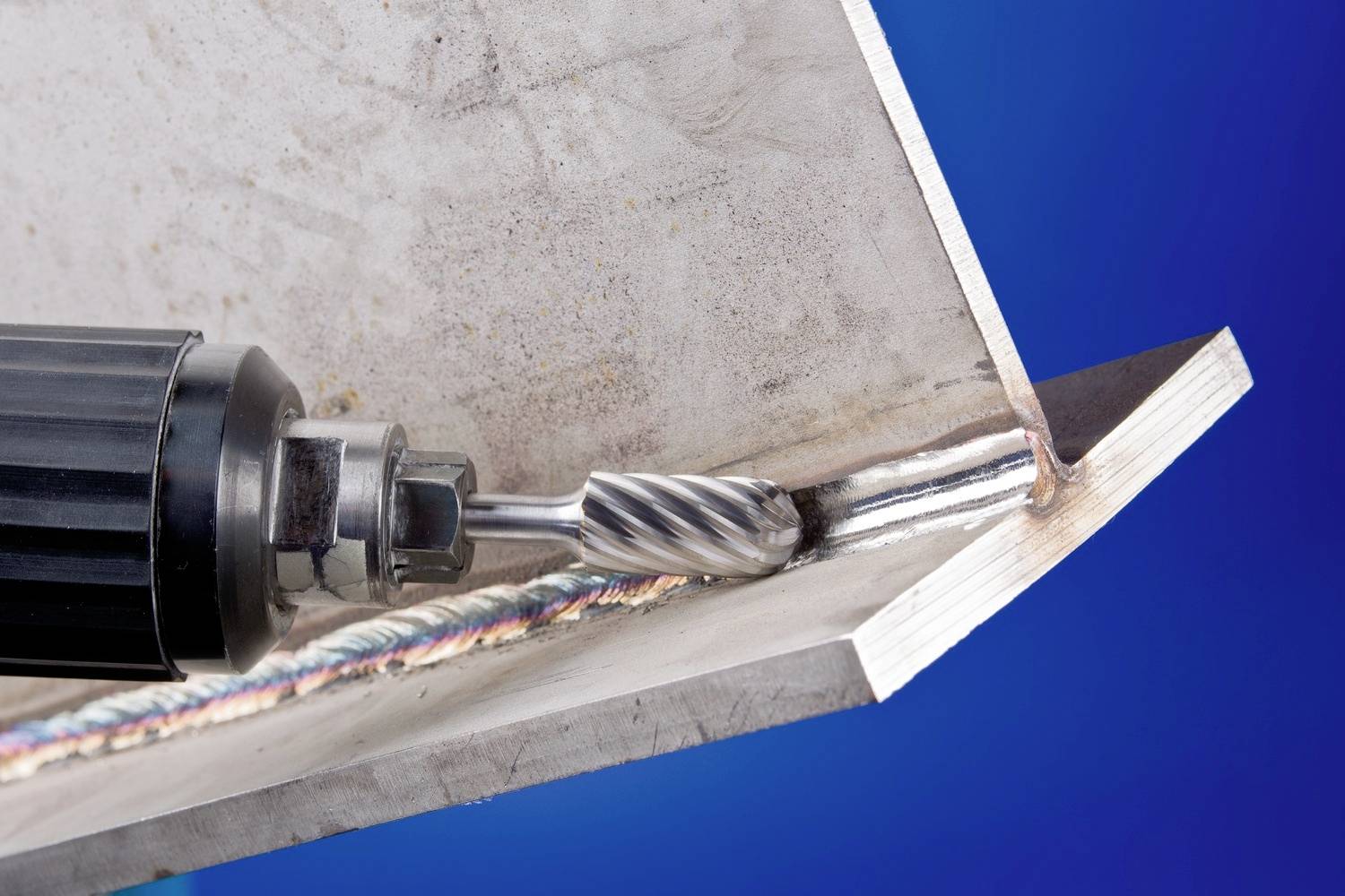 Nahaufnahme einer Metallschweißnaht. Ein elektrisches Werkzeug mit rotierender Feile bearbeitet die Naht in einem Winkel. Hintergrund in Blau.