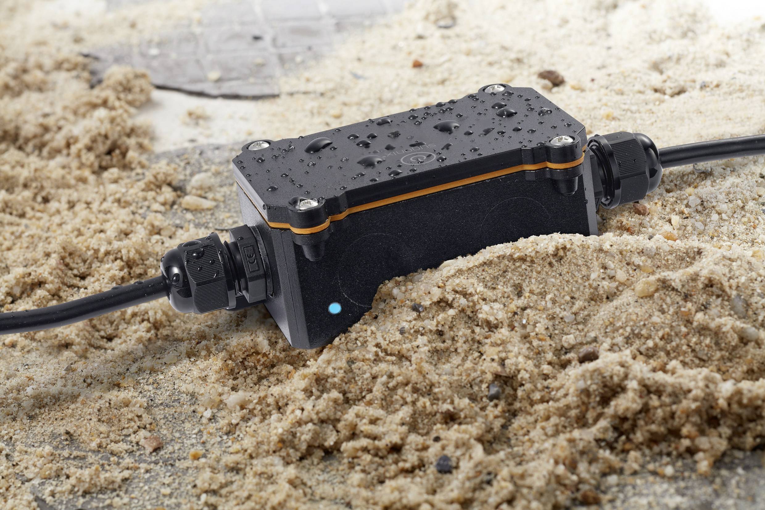 Ein wasserdichtes schwarzes elektrisches Anschlussgehäuse liegt im Sand, mit Wassertropfen und angeschlossenen Kabeln an den Enden.