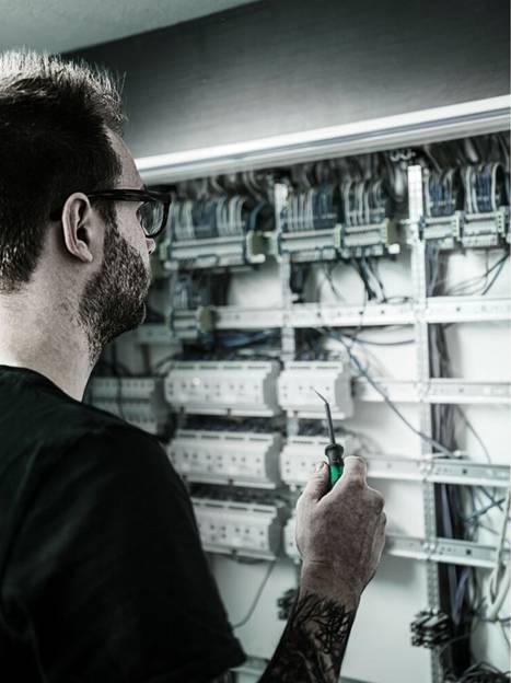 Eine Person, die einen Schraubendreher hält, untersucht eine große elektrische Schaltanlage mit zahlreichen Kabeln und Schaltern, was auf eine Wartungsaufgabe hindeutet.