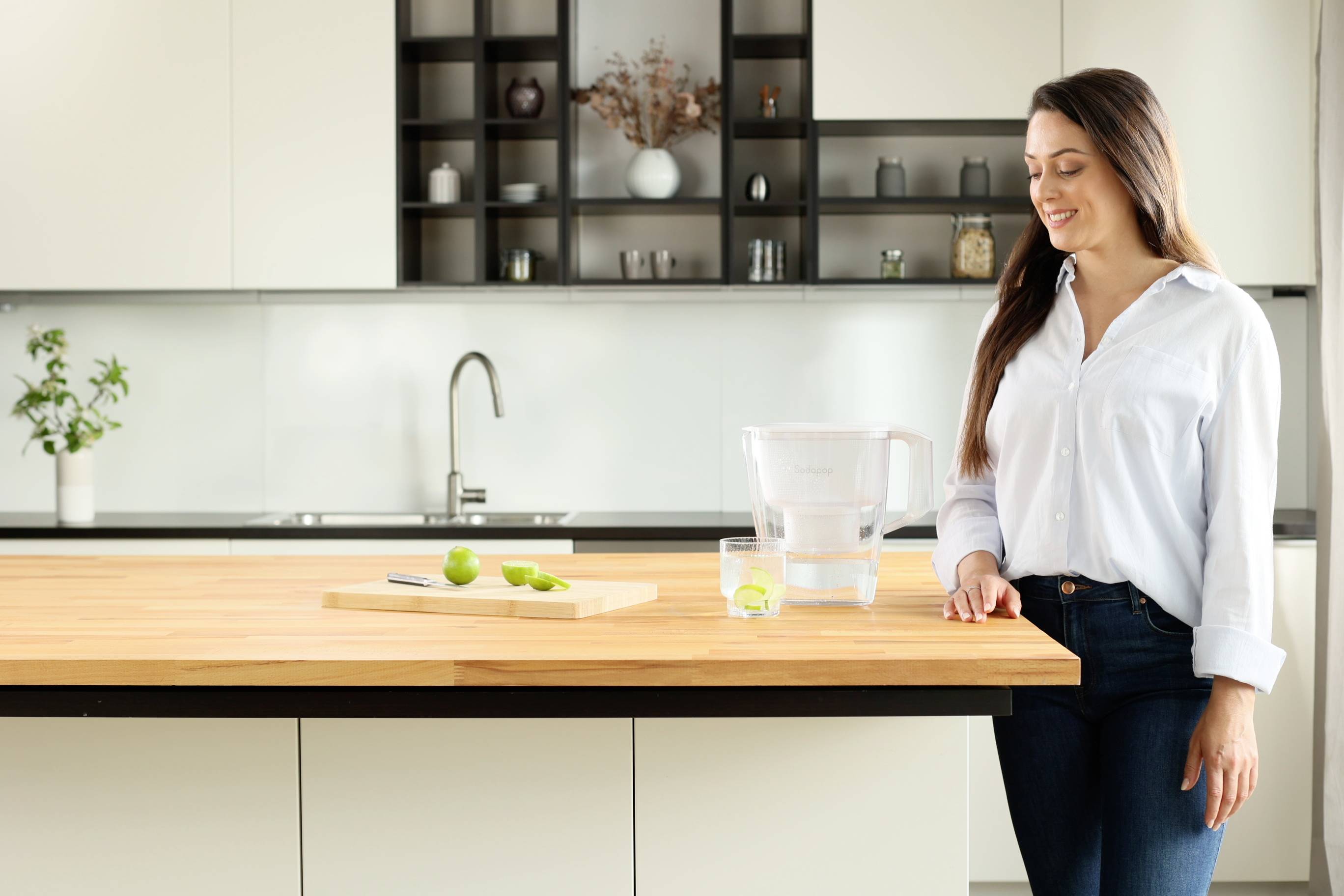 Frau steht in moderner Küche neben Wasserfilter auf Holztisch, mit Limetten und Glas. Lächelnd, entspanntes Ambiente.