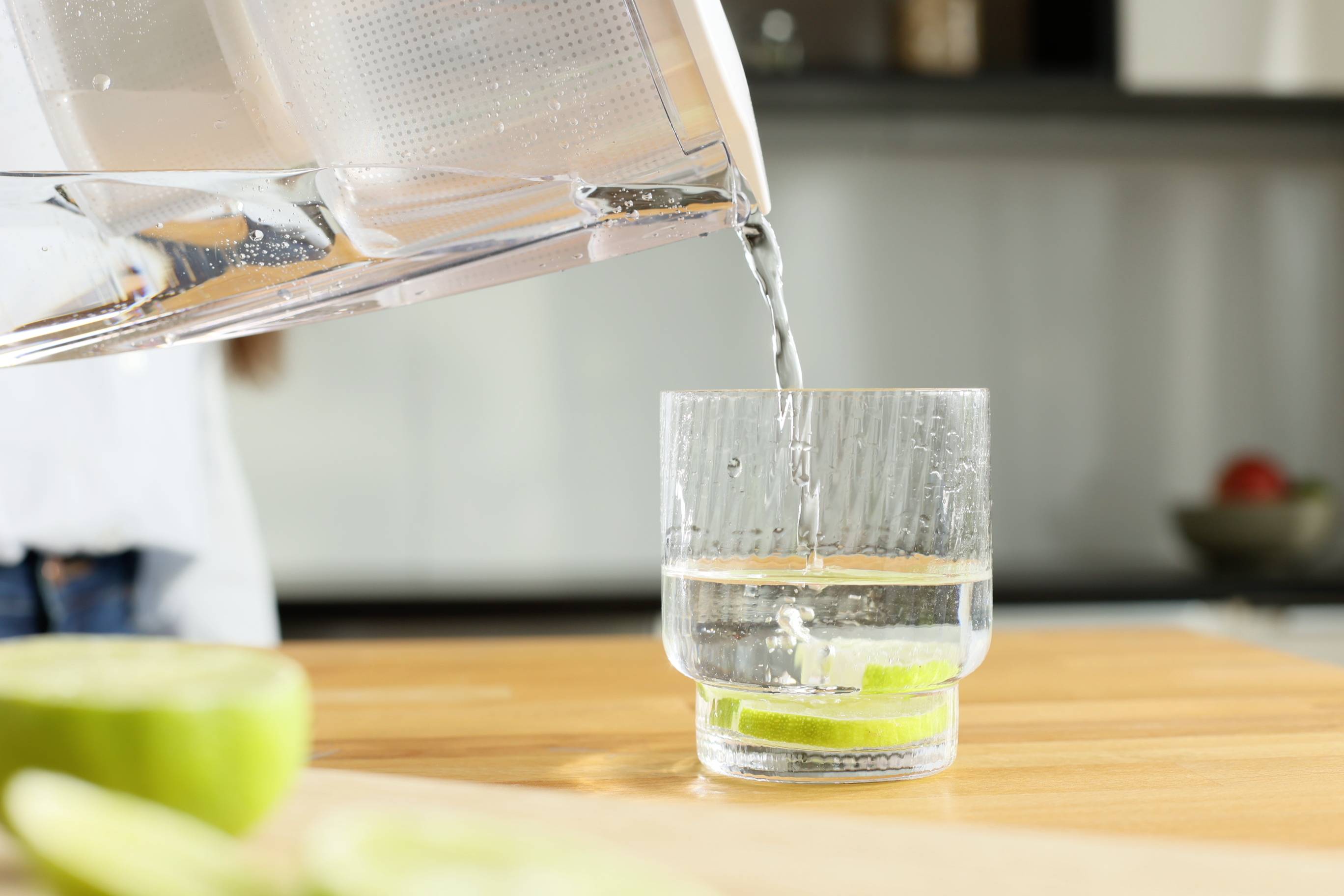 Ein Glas Wasser mit Limettenscheiben wird auf einem Holztisch gefüllt. Im Hintergrund ist eine verschwommene Küche sichtbar.