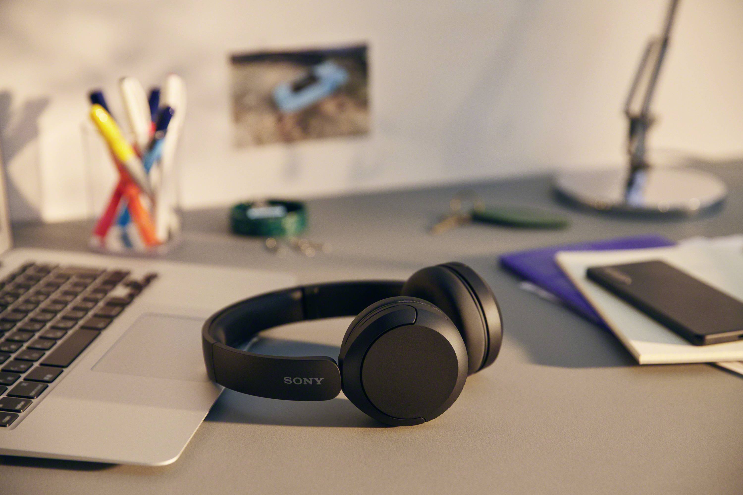 Ein Laptop und schwarze Kopfhörer liegen auf einem Schreibtisch. Daneben sind Stifte, ein Notizblock und eine Lampe zu sehen.