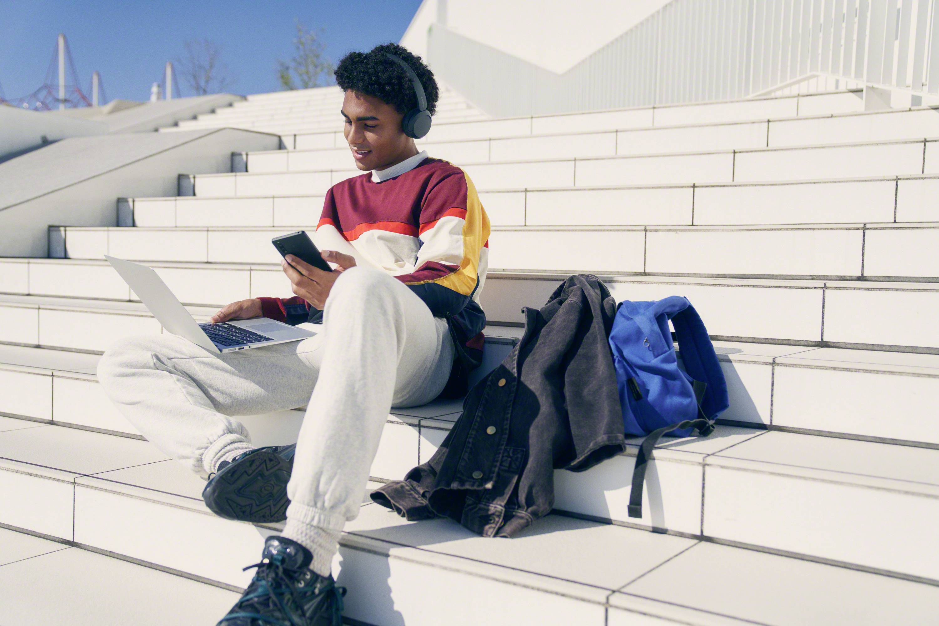 Ein junger Mann sitzt auf weißen Treppen, trägt Kopfhörer und benutzt ein Smartphone und Laptop. Neben ihm liegen eine Jacke und ein blauer Rucksack.