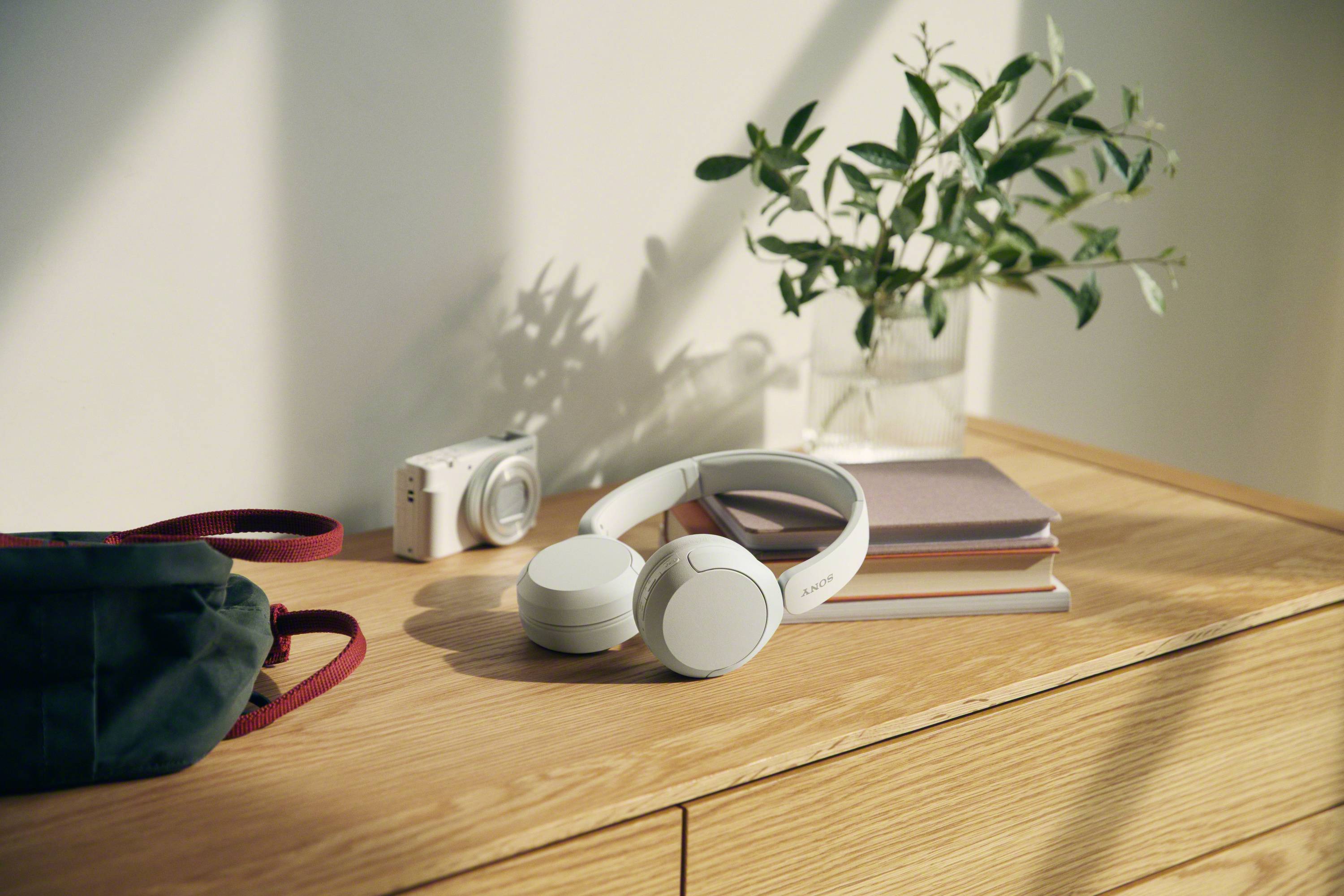 Weiße Kopfhörer auf einem Stapel Bücher auf einem Holztisch, neben einer Kamera und einer Pflanze; entspannte Atmosphäre.