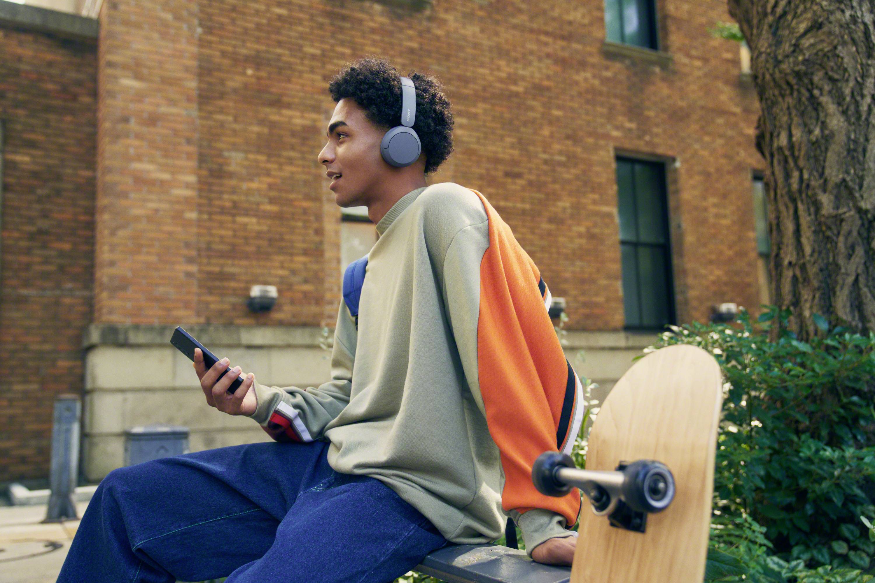 Eine Person mit Kopfhörern sitzt mit einem Smartphone und einem Skateboard auf einer Bank vor einem Gebäude.