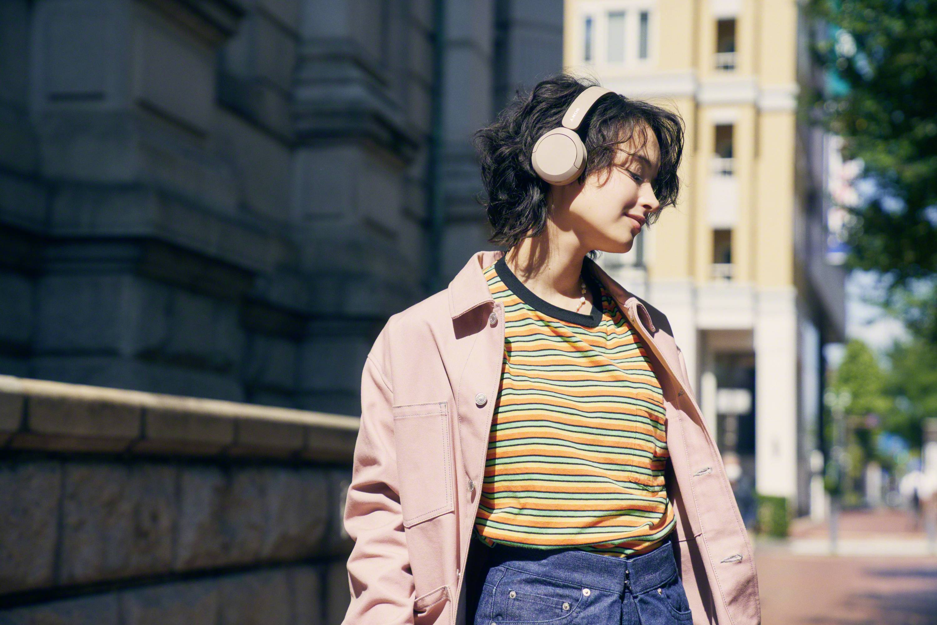 Person mit Kopfhörern, gekleidet in Streifenshirt und Jacke, spaziert an einem sonnigen Tag durch eine städtische Straße.