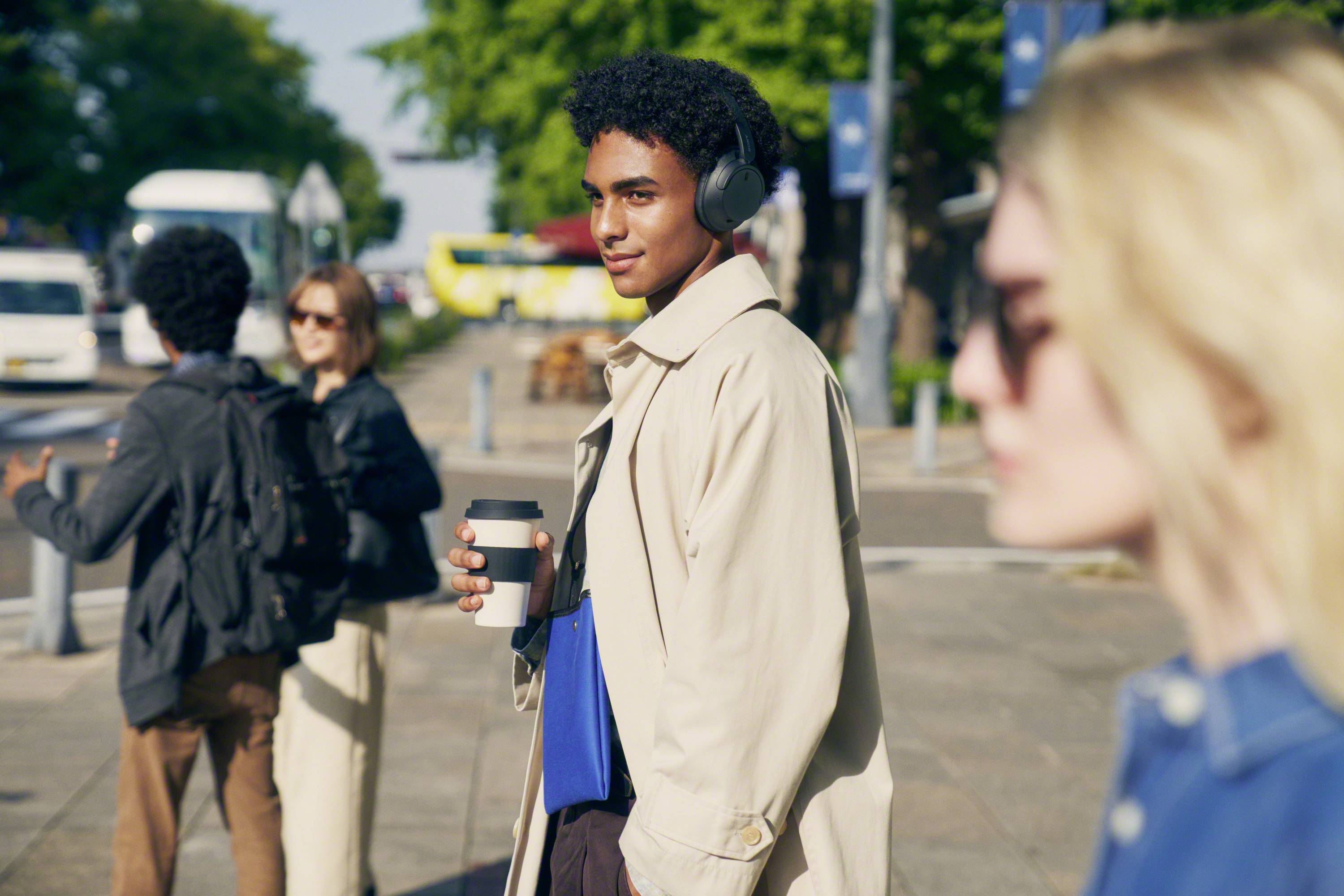 Eine Person mit Kopfhörern geht eine belebte Straße entlang, hält einen Kaffee und trägt eine Mappe. Andere Menschen sind unscharf im Hintergrund.