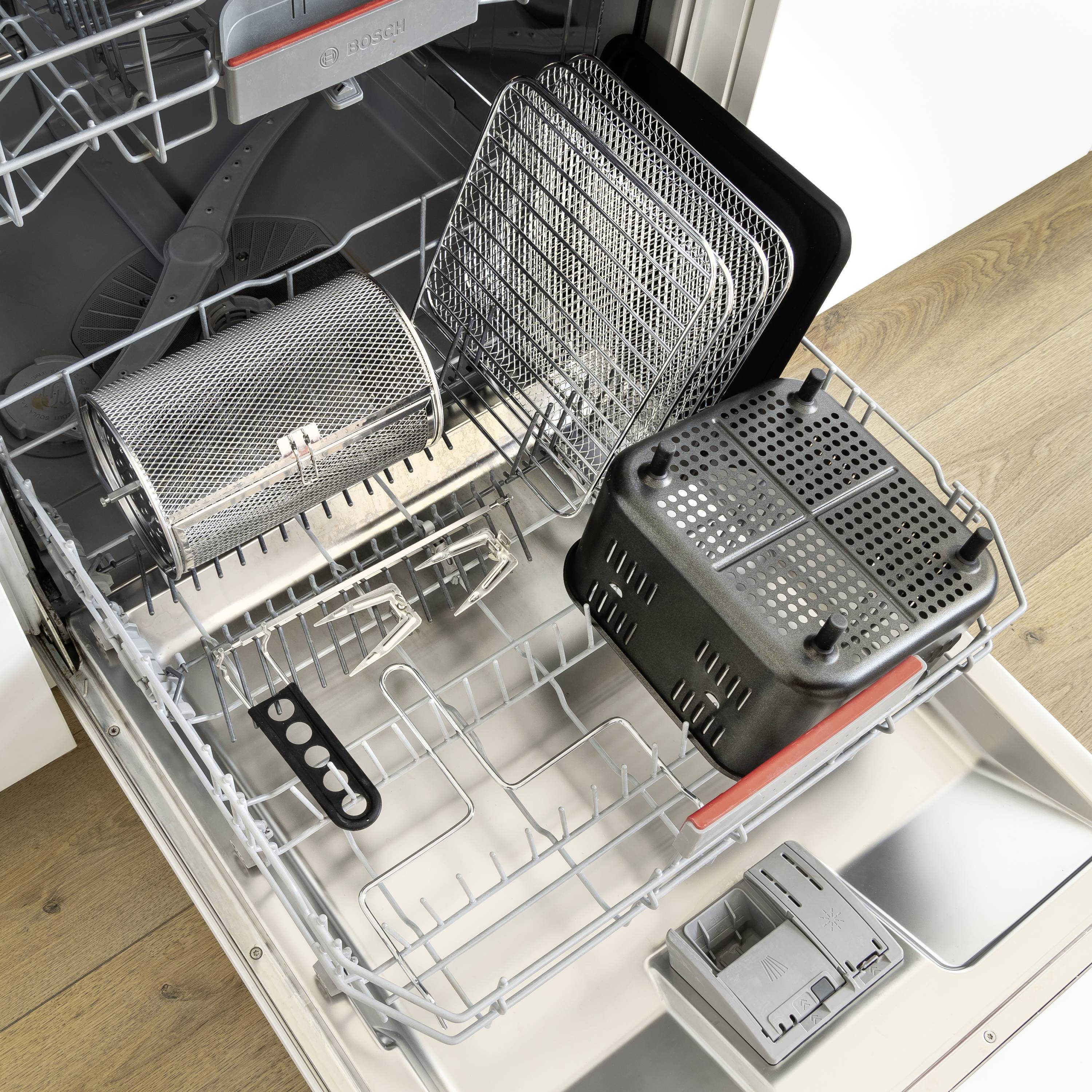 Ein geöffneter Geschirrspüler mit mehreren Metallgittern, einer Besteckablage und einem Filterkorb im unteren Fach.