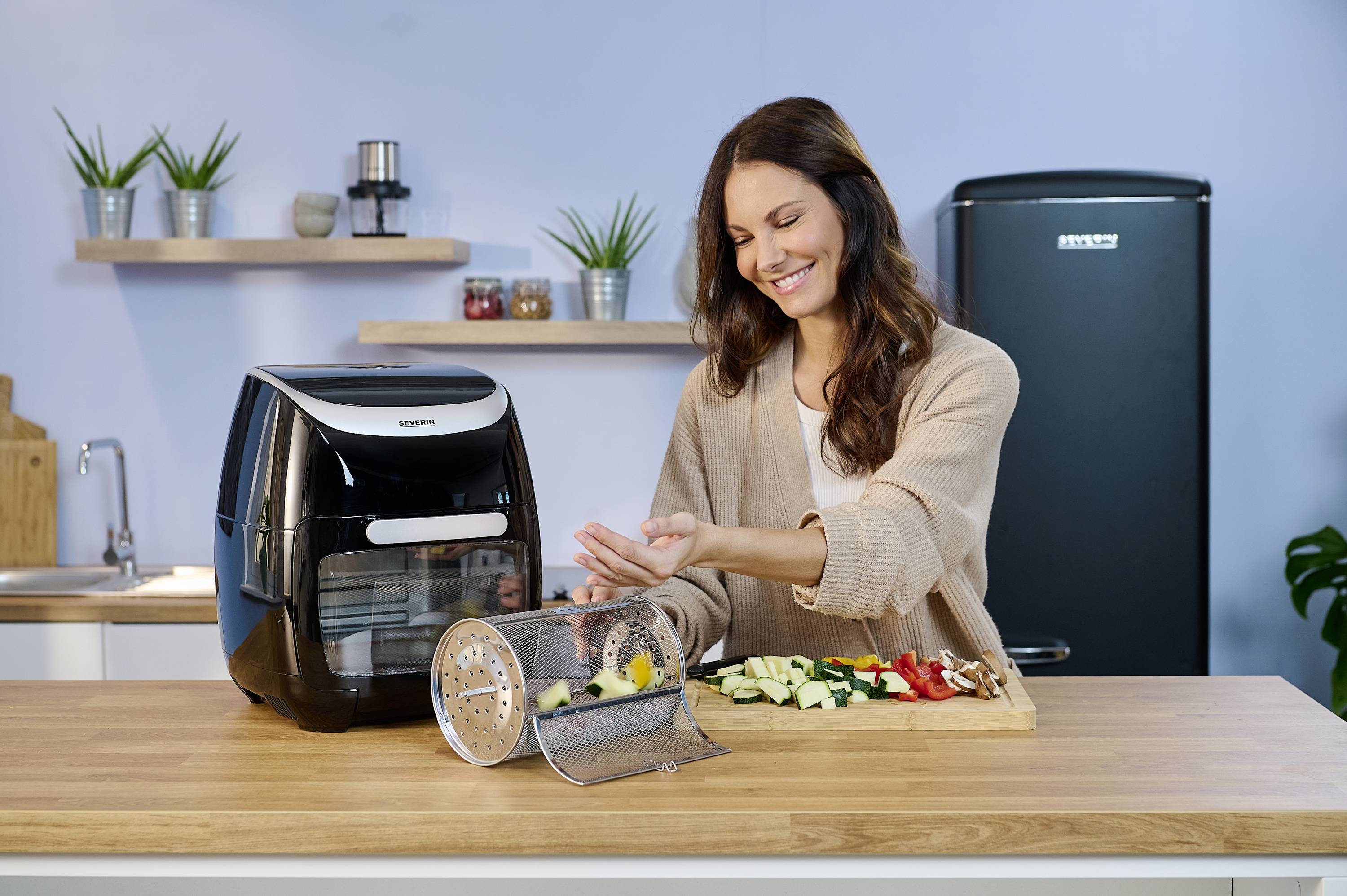 Eine Frau bereitet Gemüse in einer modernen Küche zu. Neben ihr steht eine Heißluftfritteuse auf der Arbeitsplatte. Im Hintergrund sind Küchenutensilien zu sehen.