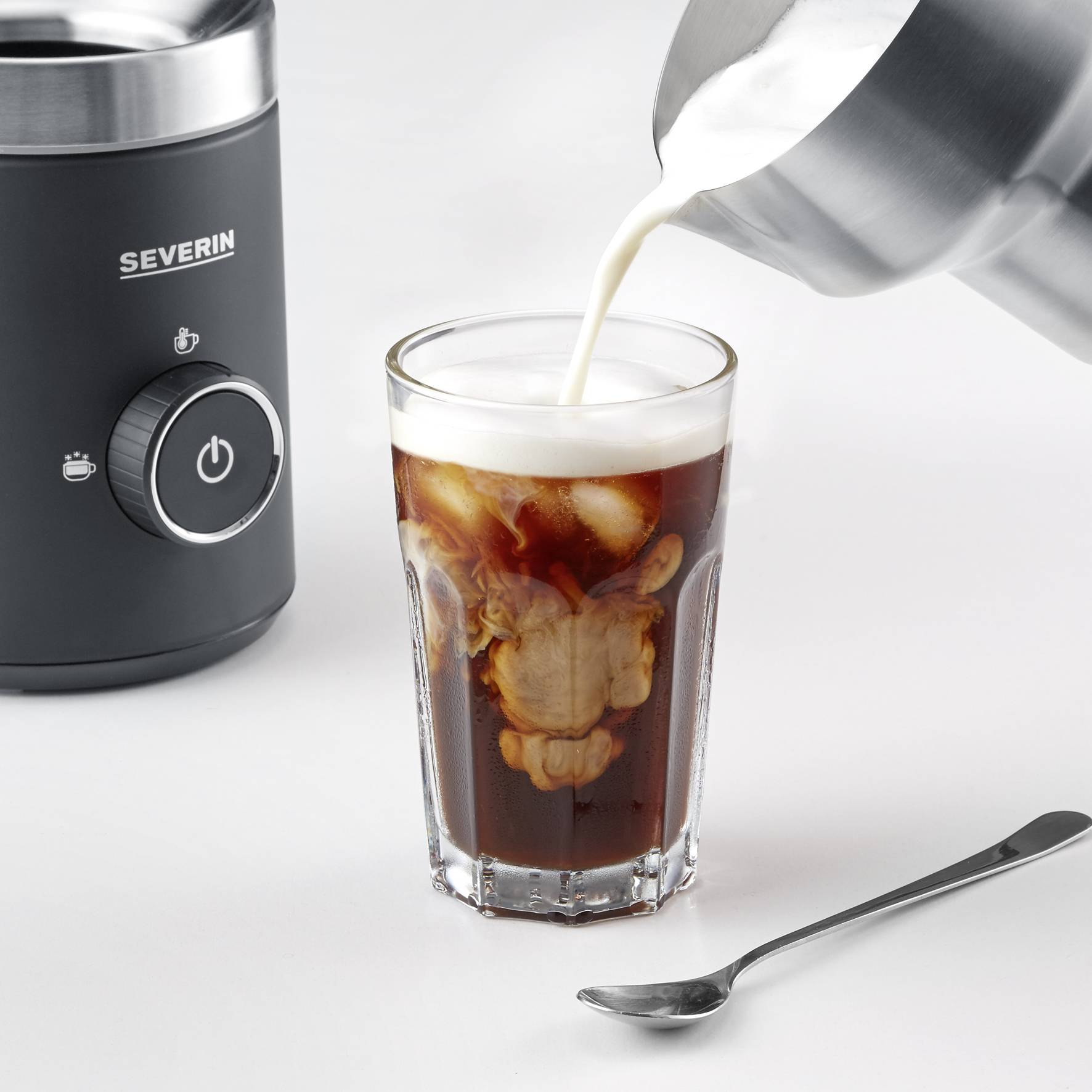 Ein Glas Eiskaffee wird mit Milch aus einem Edelstahlbehälter aufgegossen. Daneben steht ein elektrisches Gerät und ein Löffel.