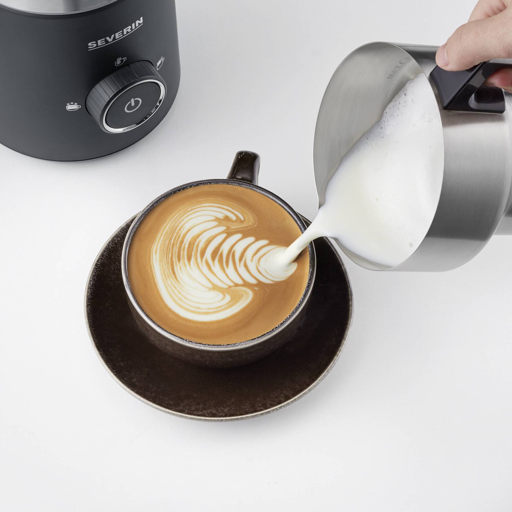 Eine braune Tasse mit Latte Art wird mit Milch aus einem Metallkrug befüllt. Im Hintergrund ist ein Milchaufschäumer zu sehen.