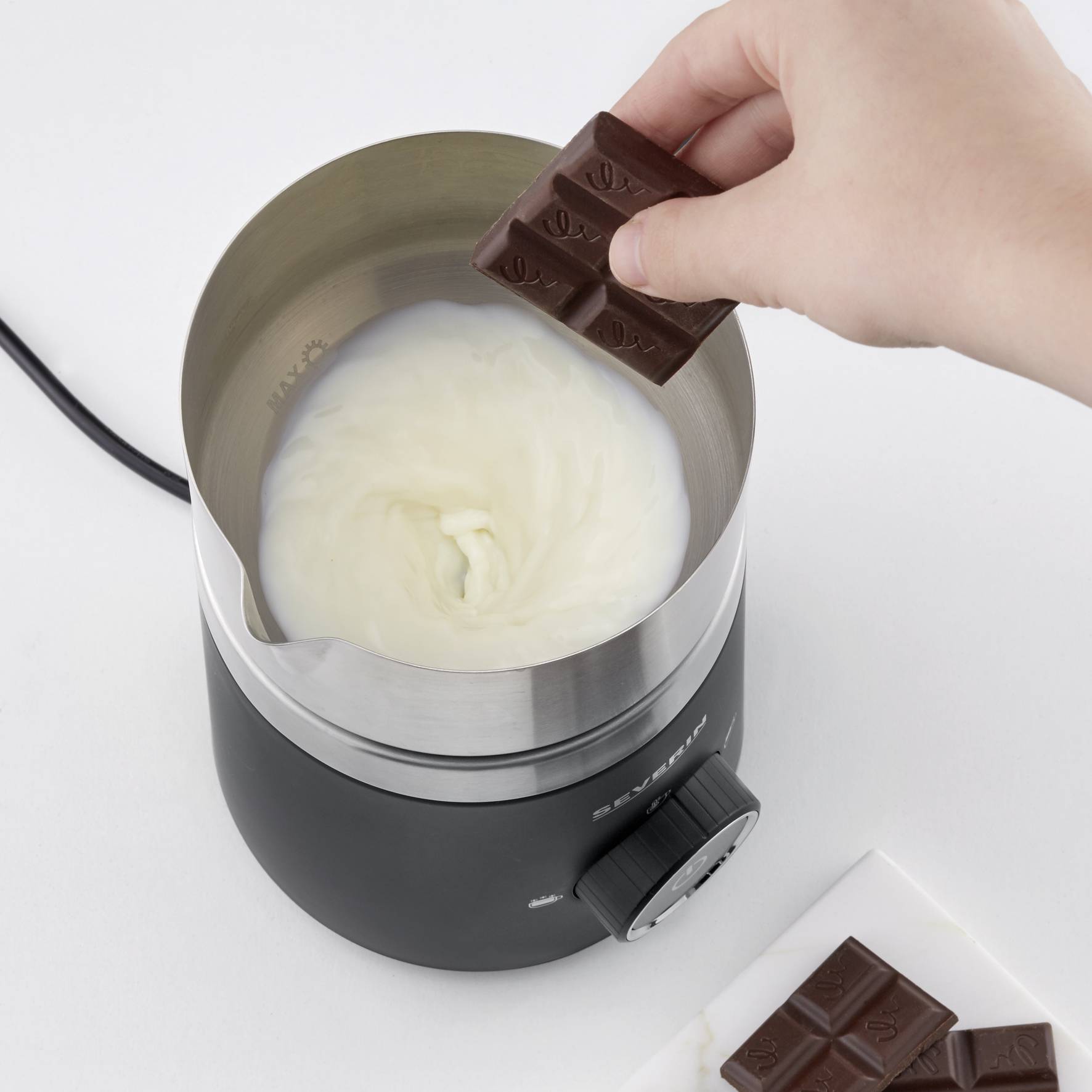 Eine Hand hält ein Schokoladenstück über einen Topf mit sich drehender Milchcreme in einem elektrischen Mixer.