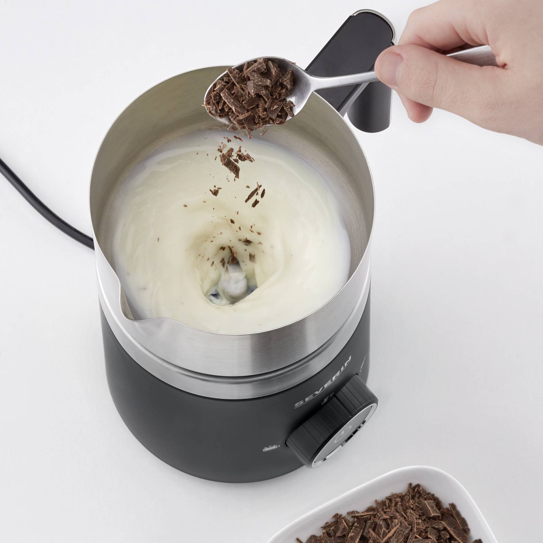 Schüssel eines elektrischen Schokoladen-Fondues. Eine Hand hält einen Löffel mit geriebener Schokolade über die erhitzte Flüssigkeit.