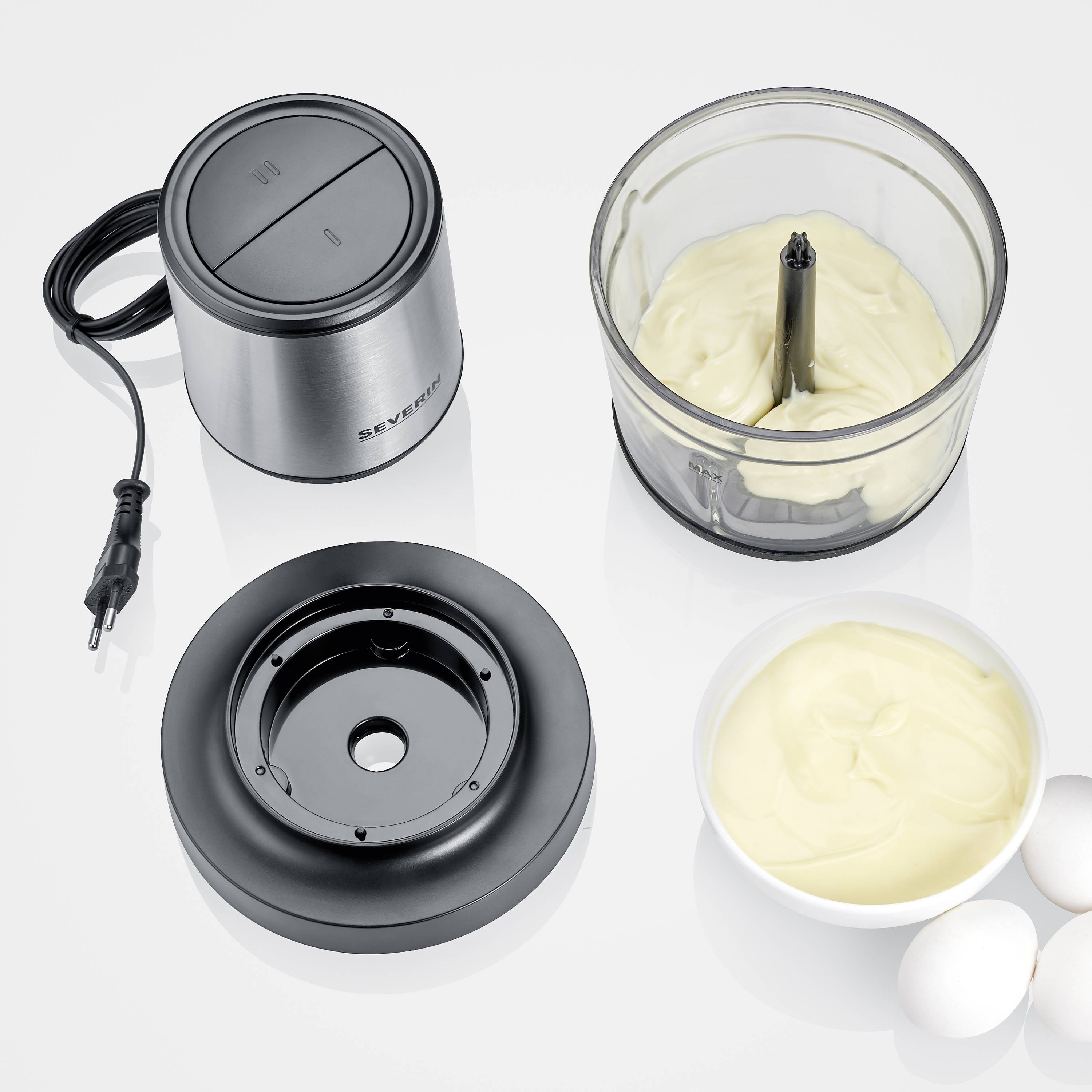 Ein Mixer mit abgenommenem Deckel, gefüllt mit gelber Creme. Daneben eine Schüssel mit Creme und zwei Eier auf einer weißen Oberfläche.