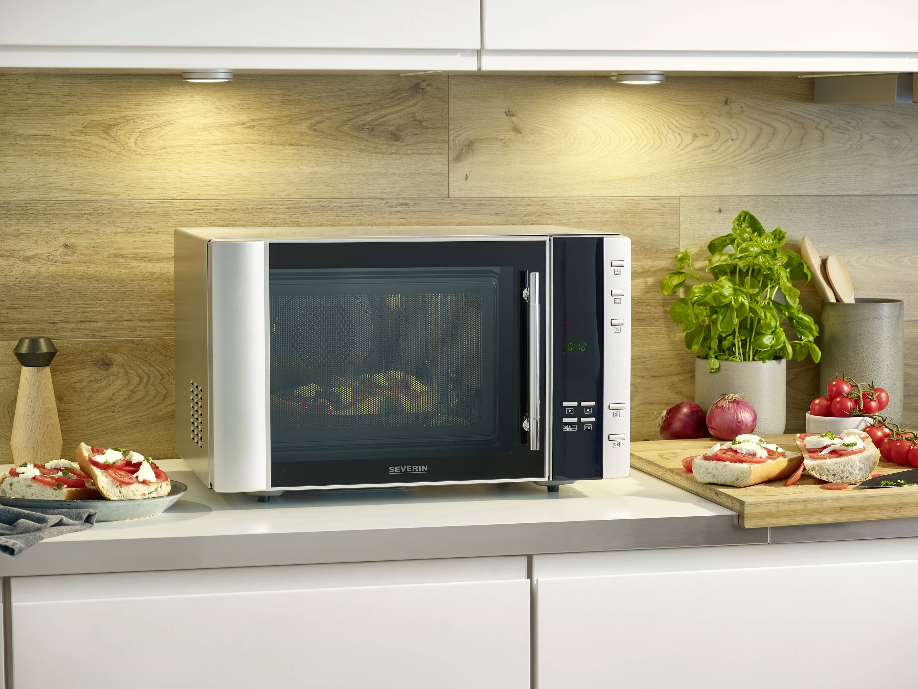 Mikrowelle auf Küchenarbeitsplatte, umgeben von Küchenutensilien und frischen Zutaten, darunter Kräuter und belegte Brote.