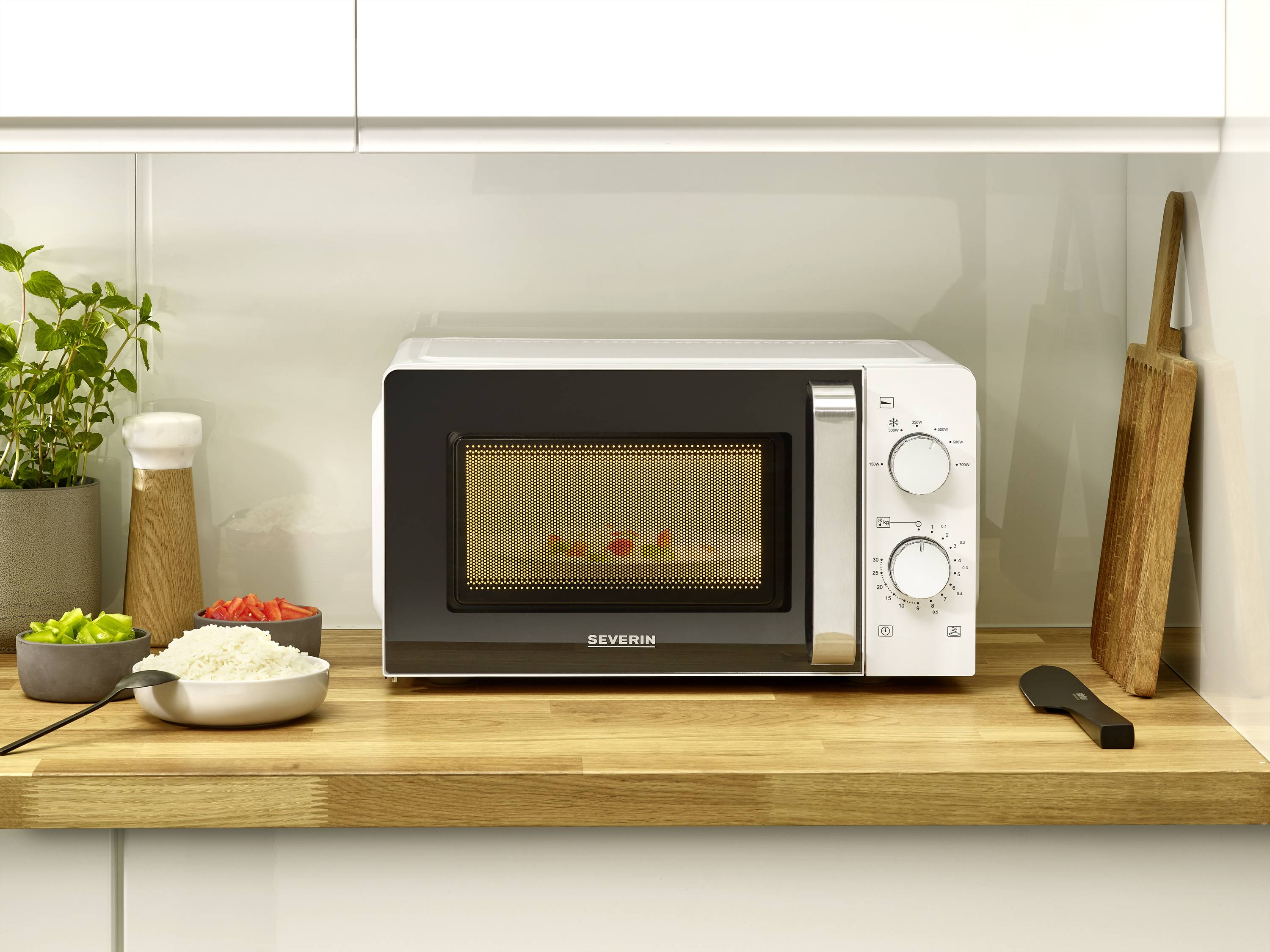 Mikrowelle auf Holzarbeitsplatte, daneben Gewürze und Geschirr; im Inneren Speisen auf einem Teller. Küche mit weißem Schrank.