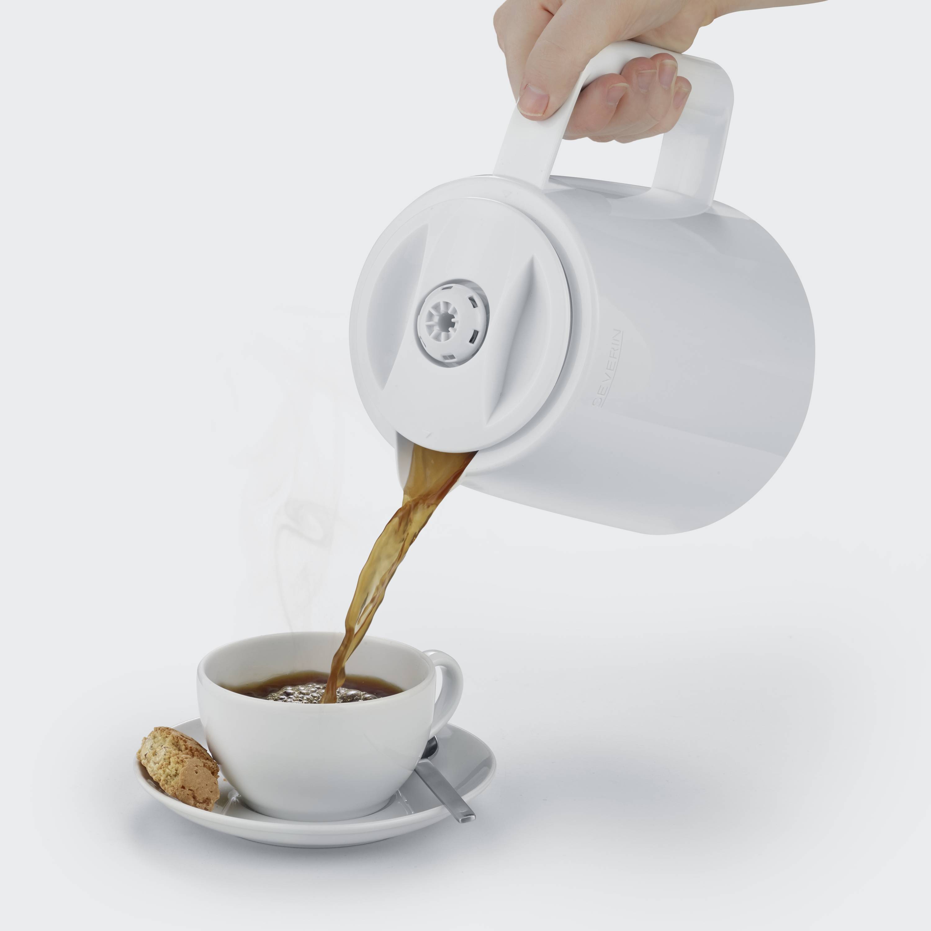 Ein weißer Wasserkocher gießt heißen Tee in eine Tasse mit Unterteller, daneben liegt ein Keks. Dampf steigt aus der Tasse auf.