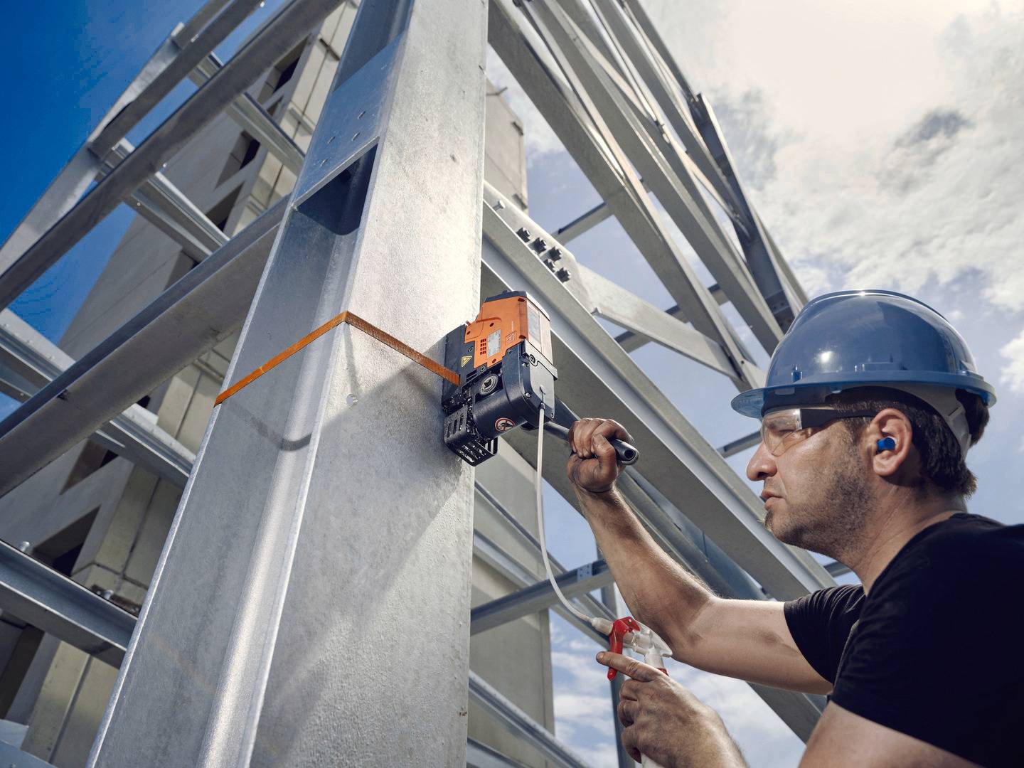 Ein Arbeiter trägt einen blauen Schutzhelm und befestigt mit einem Werkzeug ein Gerät an einer Stahlkonstruktion unter freiem Himmel.