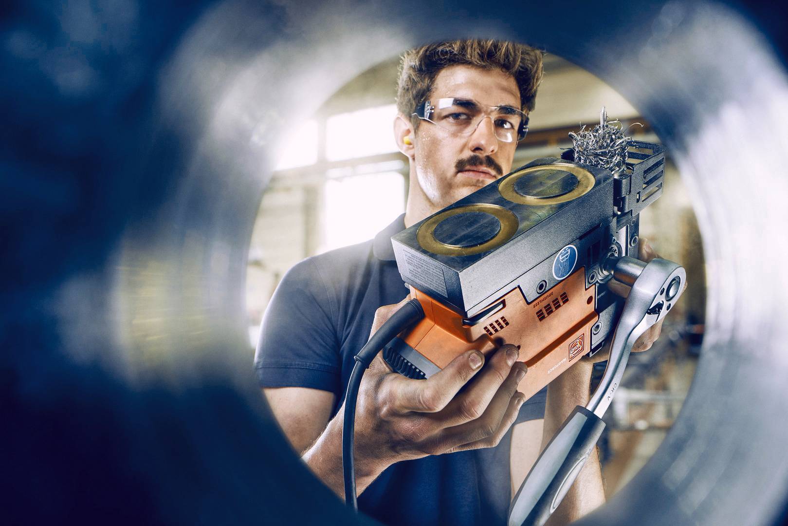 Ein Mann mit Schutzbrille hält ein orange-schwarzes Gerät, möglicherweise ein technisches Werkzeug. Er befindet sich in einer industriellen Umgebung.