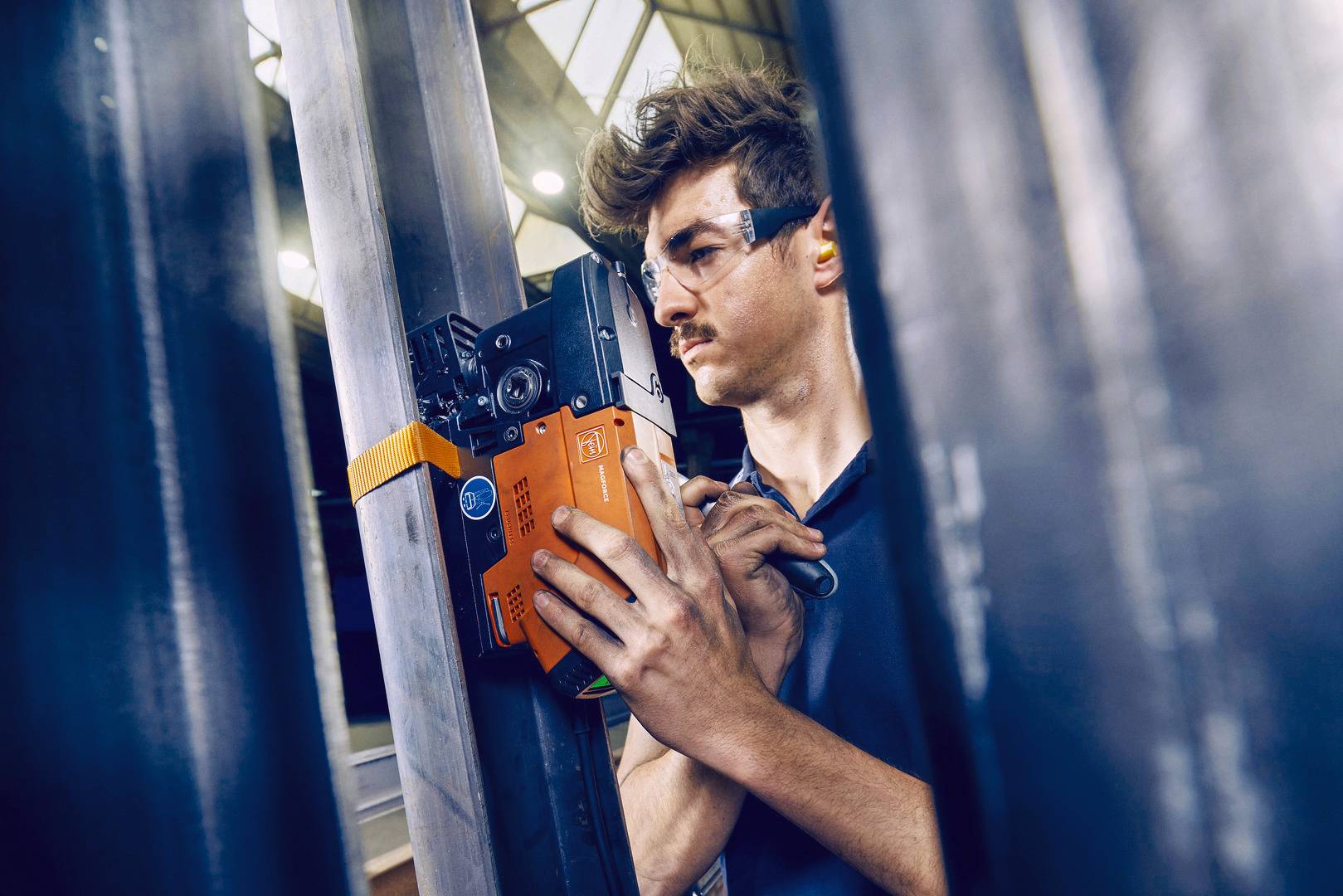 Ein Mann mit Schutzbrille überprüft mit einem Messgerät die Struktur eines Metallträgers in einem industriellen Umfeld.