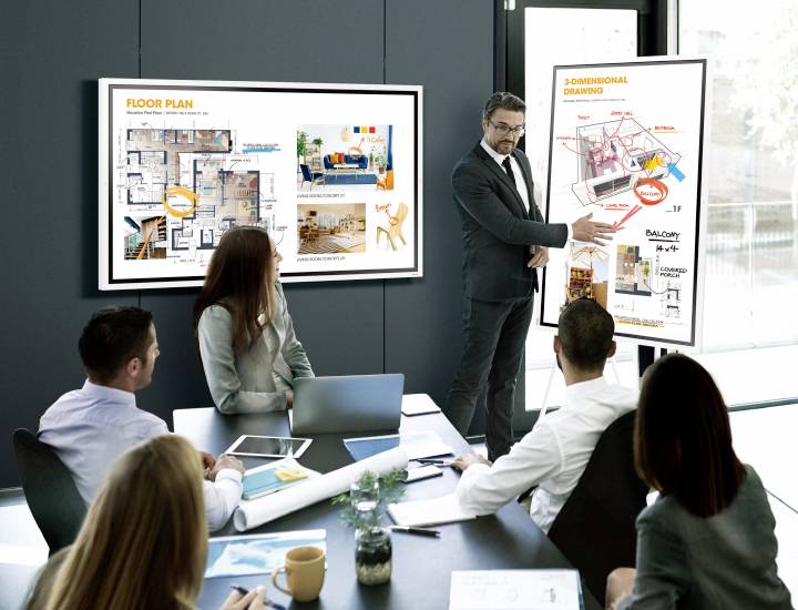 Ein Geschäftsmann präsentiert Gebäudediagramme und Grundrisse auf zwei Bildschirmen während eines Meetings mit vier Kolleginnen und Kollegen.