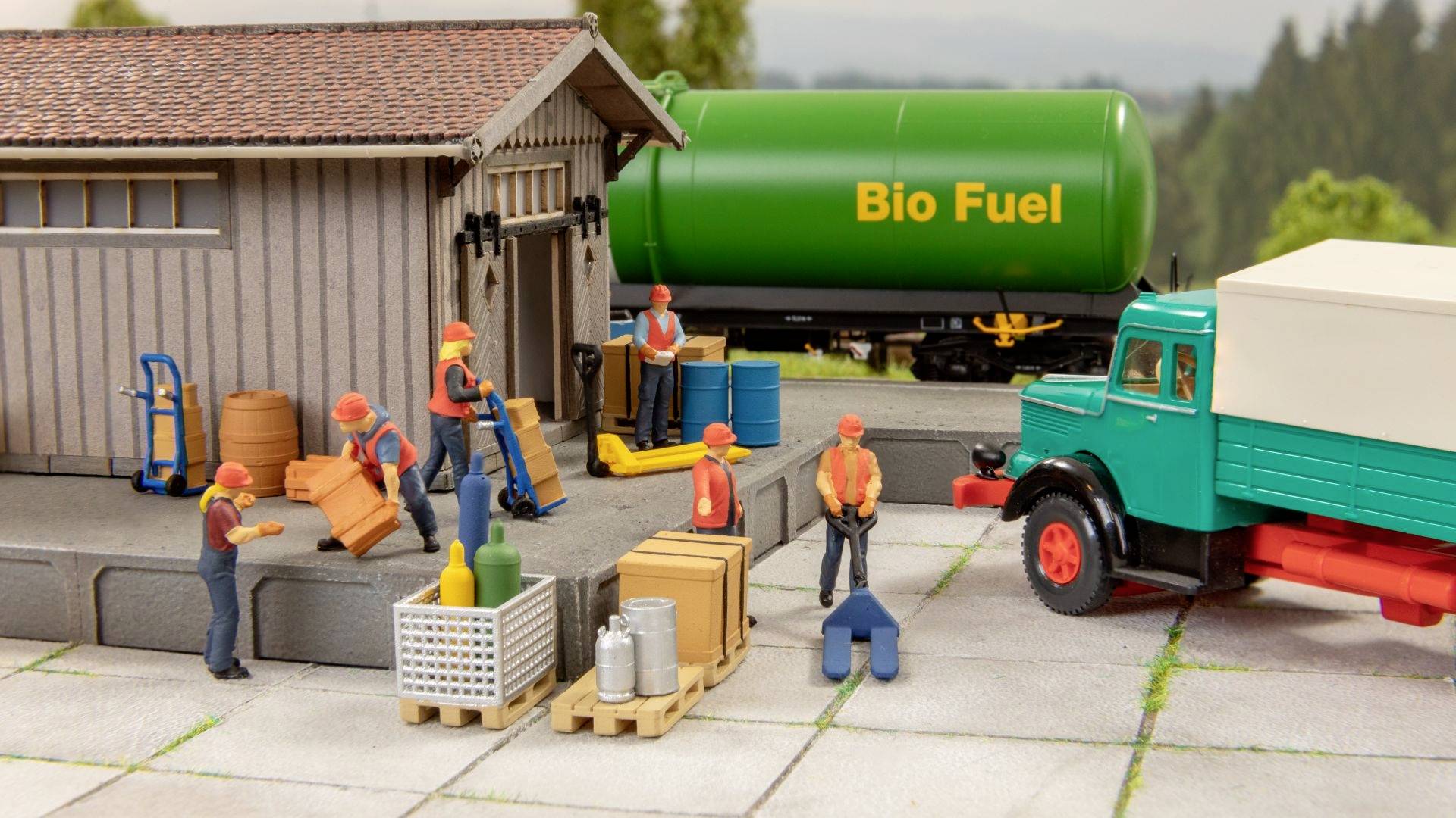 Figuren be- und entladen Fracht an einer Lagerhalle. Ein grüner Tank mit 'Bio Fuel'-Aufschrift steht im Hintergrund.