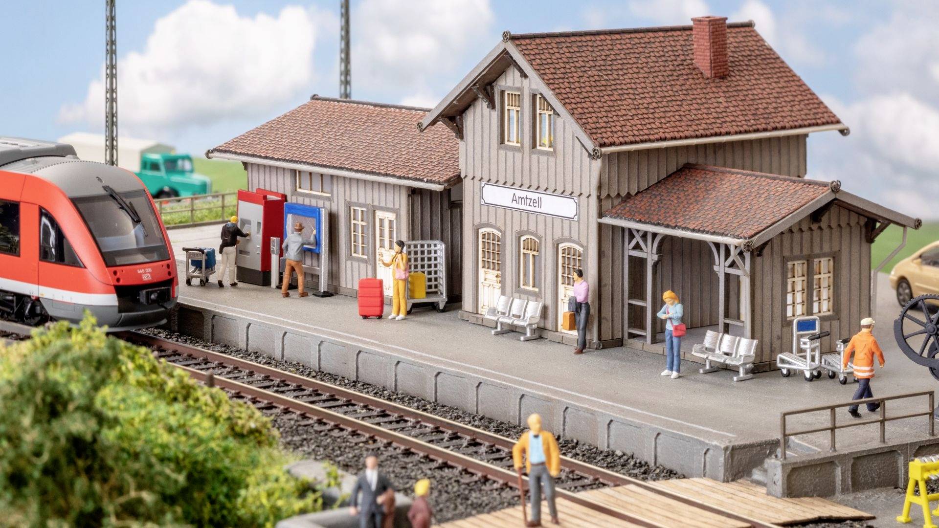 Ein Modellbahnhof mit wartenden Figuren, ein Zug im Bahnhof, und ein Schild mit der Aufschrift 'Amtszell'.