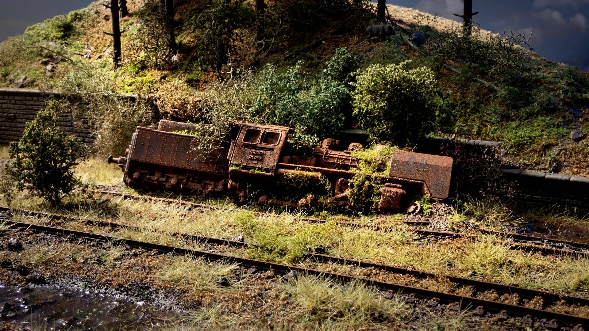 Eine verrostete, umgestürzte Lokomotive liegt neben Bahngleisen in einer bewachsenen ländlichen Umgebung.