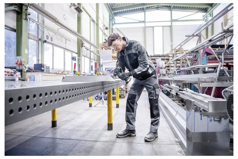 Arbeiter in Schutzausrüstung verwendet ein tragbares Werkzeug zur Inspektion oder Montage eines großen Metallbalkens in einer Industriefabrik.