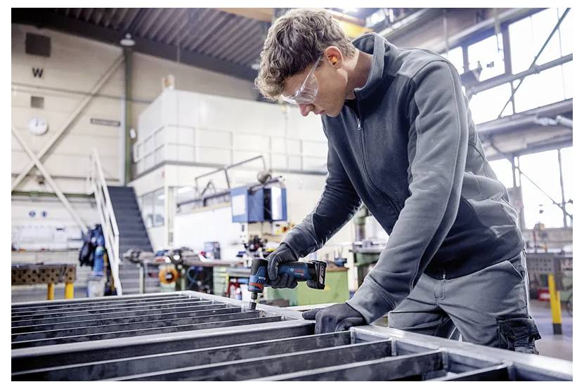 Arbeiter in Schutzbrille und Handschuhen, der einen Metallrahmen in einer Werkstatt oder Fabrik misst oder montiert.