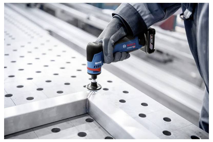 Ein Arbeiter mit Handschuhen verwendet ein schnurloses Elektrowerkzeug von Bosch, um ein Metallblech auf einem perforierten industriellen Arbeitstisch zu befestigen.