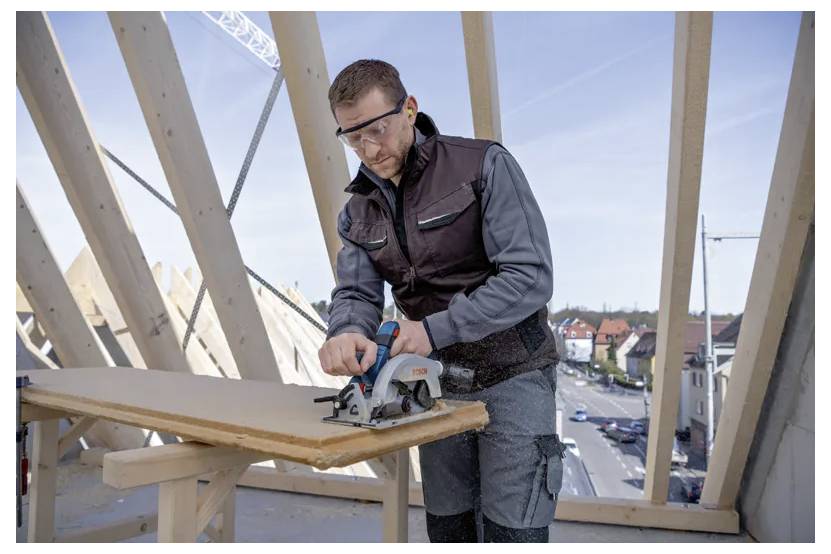 Eine Person in Schutzausrüstung bedient eine Kreissäge an einem Stück Holz auf einer Baustelle mit einer Stadt im Hintergrund.