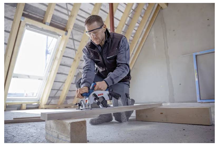 Eine Person mit Schutzbrille verwendet eine Kreissäge, um ein Holzbrett in einem unfertig ausgebauten Dachboden zu schneiden, während Licht durch ein Fenster fällt.